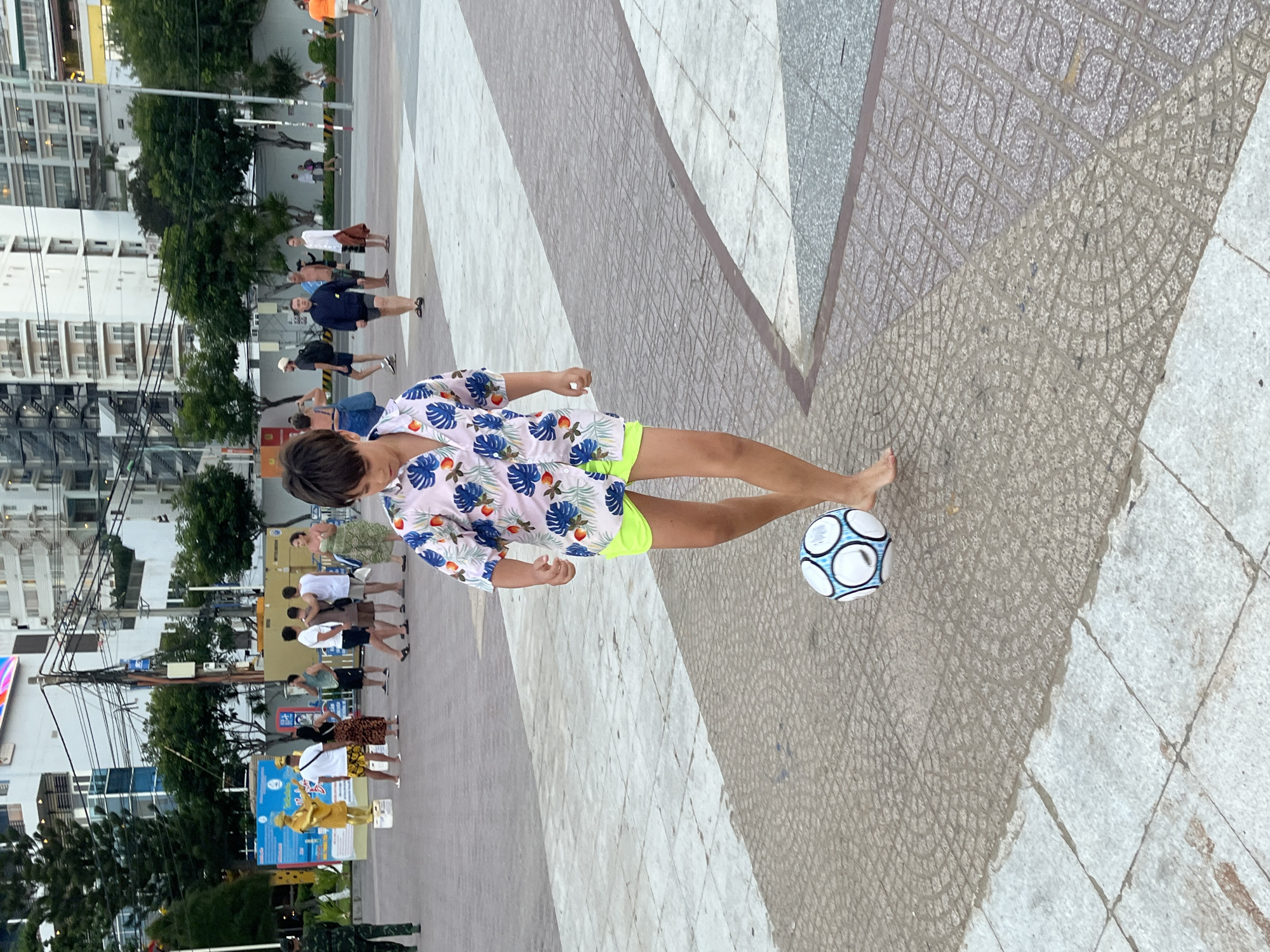 A young male wearing a floral shirt and neon green shorts is playing soccer on a paved plaza. The plaza is made of interlocking hexagonal and square tiles. In the background, there are several buildings, trees, and other people walking around. To the left of the young male, there are two gold statues. The scene appears to be in Nha Trang, Vietnam, a coastal city known for its beaches and resorts.