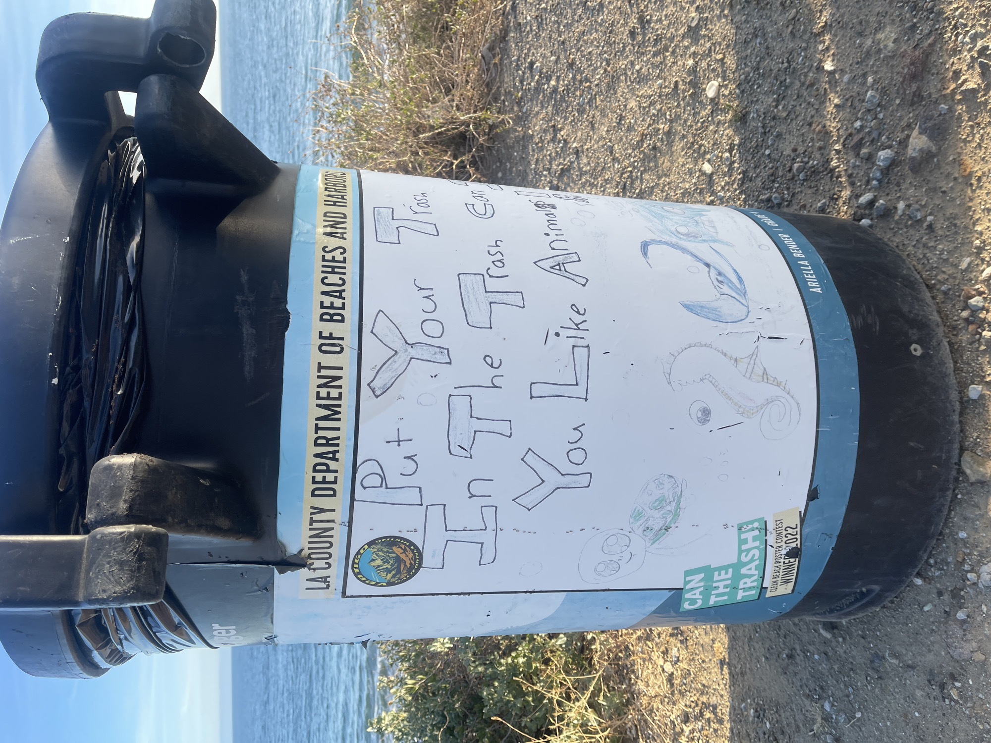 The image shows a black trash can with a white poster wrapped around it. The poster is titled "Put Your Trash In The Trash Can You Like Animals." The poster also has drawings of a seahorse, turtle and other marine life. The trash can is located near the beach in Malibu, California, United States of America. The trash can belongs to the LA County Department of Beaches and Harbors. The poster on the trash can is from the Clean Beach Poster Contest and the winner was Ariella Bender/Grade.