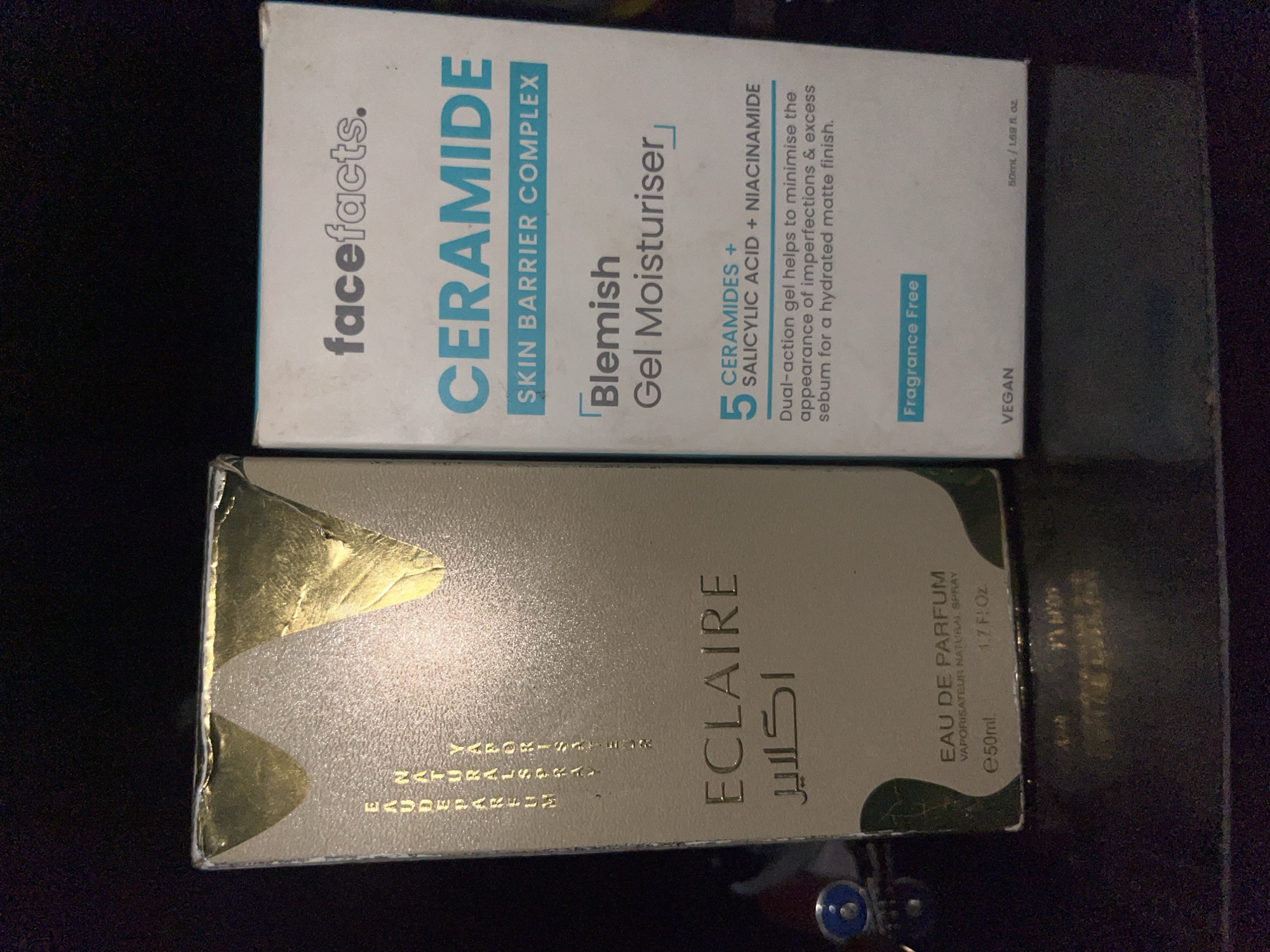 The image displays two product boxes placed on a dark, textured surface, likely a table. The box on the left is a light beige color with metallic gold accents. It features text in gold, including "EAU DE PARFUM" and "ECLAIRE" in large font, with smaller text above and below. The box on the right is white with teal and gray text. It displays the brand "facefacts." prominently at the top, followed by "CERAMIDE SKIN BARRIER COMPLEX" and "Blemish Gel Moisturiser." Below this, there is information about "5 CERAMIDES + SALICYLIC ACID + NIACINAMIDE" and a description of the product's benefits. A "Fragrance Free" label is visible near the bottom. The context of Bukuru, Nigeria is not inferable from the visual information in the image.