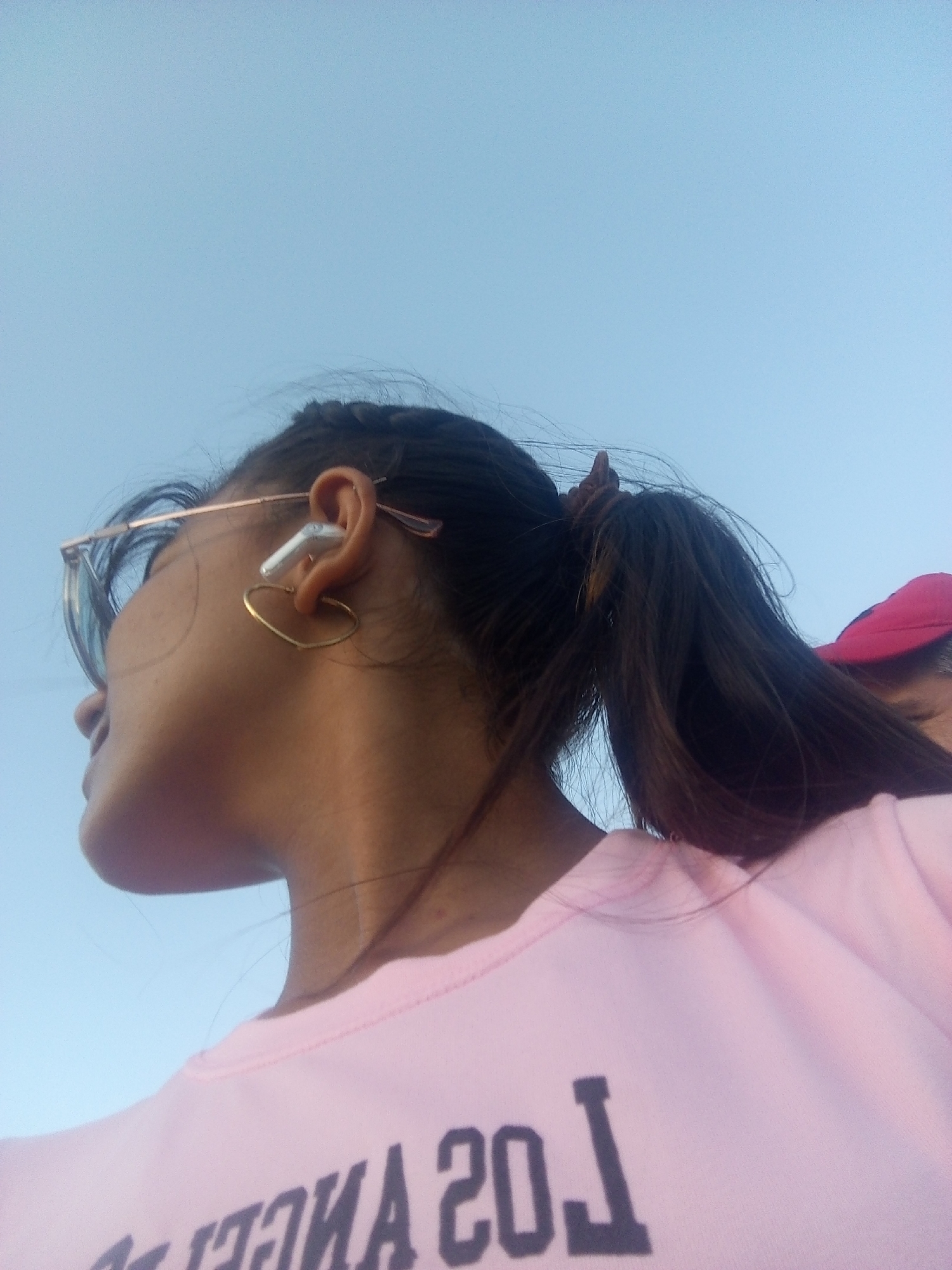 A low-angle shot shows a person with dark hair pulled back in a ponytail, wearing a pink shirt with "Los Angeles" printed in dark block letters. They are wearing gold-rimmed glasses and a heart-shaped earring in their left ear, along with a white wireless earbud. The background is a clear, light blue sky. Part of another person wearing a red hat is visible in the upper right corner. The location and city are unknown.
