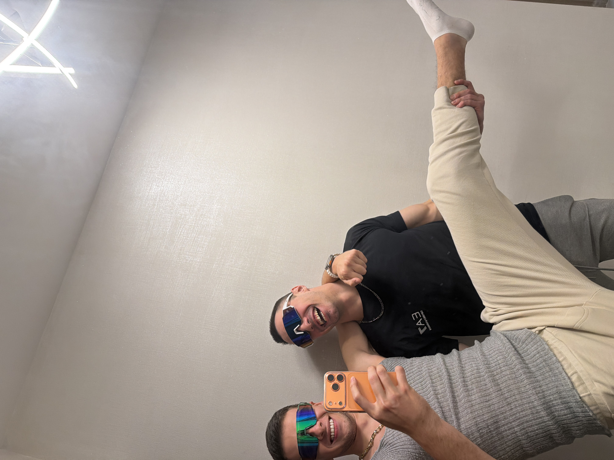An image captures two young men posing humorously for what appears to be a mirror selfie. The photo is oriented ninety degrees counter-clockwise from a standard upright view.
The man closer to the viewer, wearing a grey ribbed t-shirt and a gold-colored chain, holds an orange smartphone with multiple camera lenses, indicating he is taking the picture. He smiles broadly, showing teeth, and wears reflective, iridescent sports sunglasses.
Behind him and slightly to his right, another man in a black t-shirt and light-colored pants performs a high leg lift. He grasps his own raised left ankle with his right hand, extending his leg upwards. He also wears similar reflective sports sunglasses and smiles widely, revealing his teeth. A silver-colored chain and a watch are visible on him. The text "EMPORIO ARMANI" is partially visible on his black t-shirt.
Both men are looking directly into the camera/mirror. The background is a plain, light-colored wall with a subtle textured or possibly glittery finish. In the top left corner (when viewed upright), a modern, angular light fixture with white bars emits a bright glow. The overall mood is playful and jovial.