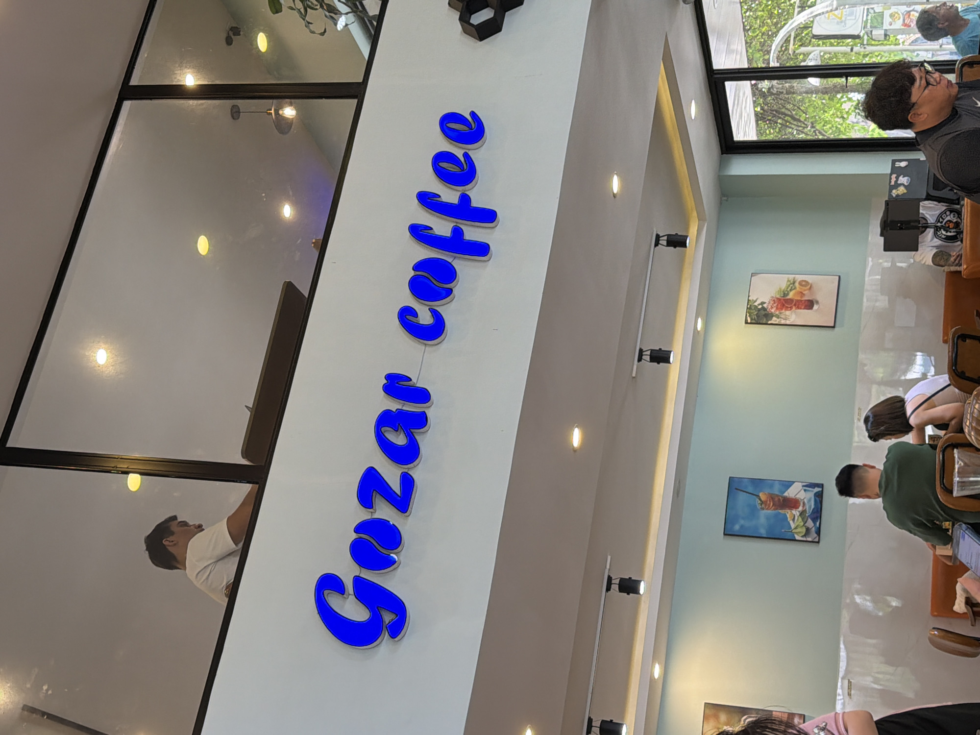 The image depicts the interior of a café in Đà Nẵng, Vietnam, identified by the blue "Gozar coffee" sign prominently displayed. The café has a modern design with a high ceiling, large windows, and recessed lighting. Several customers are visible inside, seated at tables, engaged in conversations and activities like using laptops. The café walls are decorated with framed art.