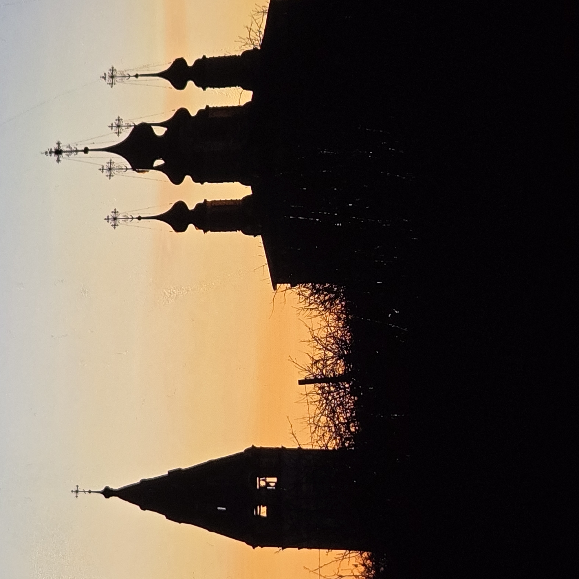 The image shows a silhouette of a bell tower and several onion domes of a church against a sunset sky. The sky transitions from pale blue at the top to orange at the horizon. The bell tower, on the left, is tall and slender with a pointed top adorned with a cross. It has visible openings that suggest it is a bell tower. To the right, the church features multiple domes of varying sizes, also topped with crosses. The foreground is filled with the dark silhouettes of bare trees and branches, suggesting a winter or early spring setting. The scene evokes a serene and historical atmosphere, typical of Russian Orthodox architecture. Based on the distinct architectural style of the bell tower and the church domes, this scene is likely located in the city of Vladimir, Russia, which is renowned for its ancient religious structures.