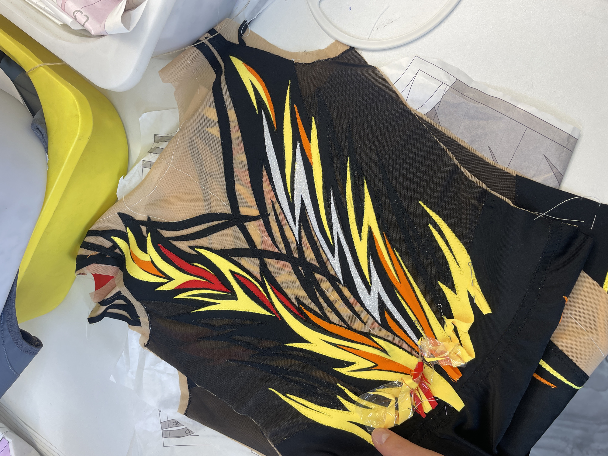 The image shows a close-up view of a partially constructed black leotard with a flame-like design in yellow, orange, red, and silver. The leotard appears to be made of mesh fabric in some areas. A sewing pattern is visible underneath the leotard. A person's hand, partially visible at the bottom left, is holding the garment and appears to be working on it, with pins and small pieces of fabric attached. In the background, a yellow plastic object, a grey fabric-like object, and a white container are visible, suggesting a sewing or crafting environment. There is no indication of the location being in Saint Petersburg, Russia.