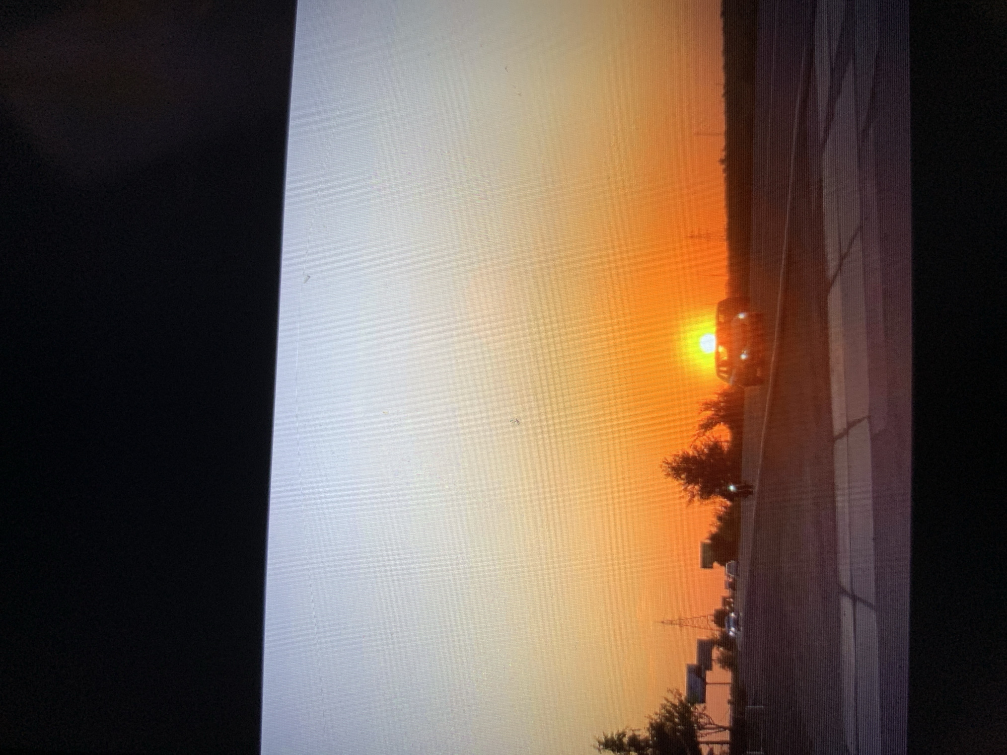 The image captures a sunset over a road in Lymanka, Ukraine. The sky transitions from a pale blue at the top to a vibrant orange and yellow near the horizon, with the sun prominently visible, partially obscured by a car. Several vehicles are on the road, including a car with its headlights on and a motorcycle. Trees and power lines dot the landscape along the roadside. The foreground shows a paved road with distinct lines.