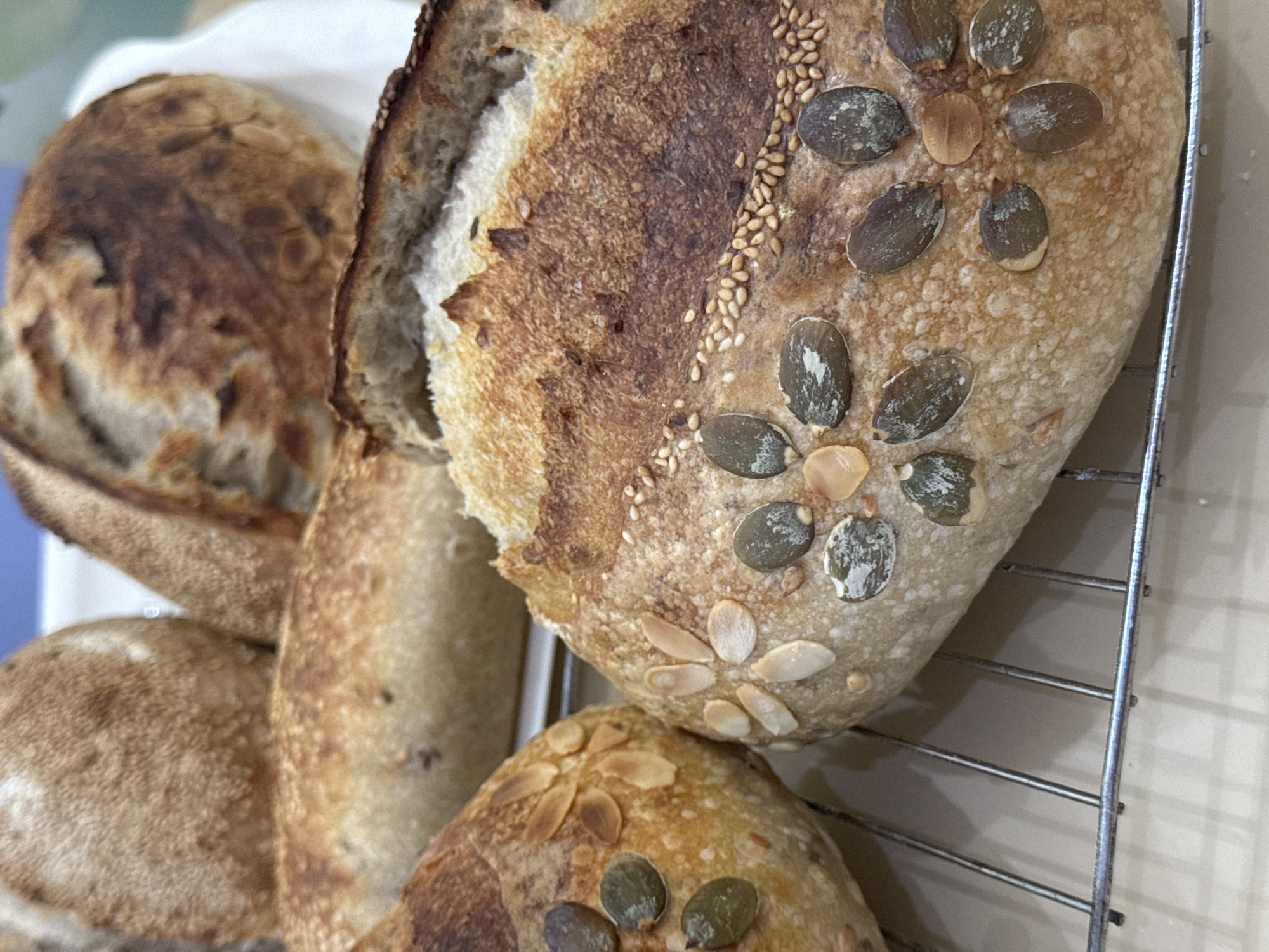 The image shows several loaves of bread cooling on a wire rack. One loaf is sliced, revealing its crumb. The bread is decorated with pumpkin seeds, sesame seeds, and almond slivers. The environment is a kitchen. The location information is not discernible from the image.