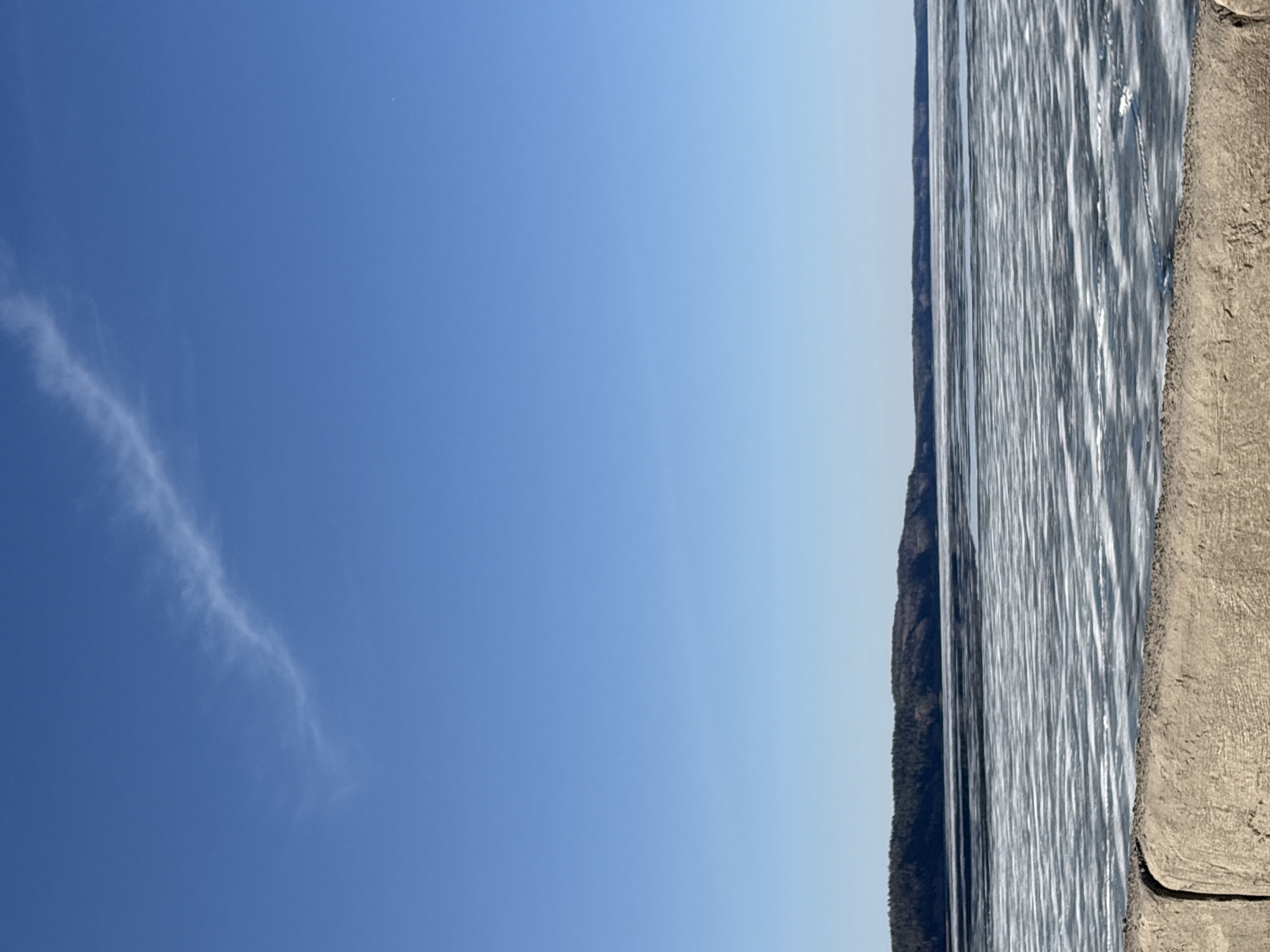 The image depicts a serene natural landscape, most likely during the daytime under clear weather conditions. The scene is dominated by a vast expanse of bright blue sky, occupying the upper two-thirds of the frame. The sky exhibits a gradient, appearing a deeper blue towards the upper left and gradually lightening towards the horizon on the right. A single, thin, wispy cloud trails across the upper left quadrant of the sky.
Below the sky, a wide body of water stretches across the midground. The water's surface is highly reflective, showing numerous vertical streaks of light and shadow, suggesting ripples or sun glare on a calm day. Across the water, a dark, forested landmass or distant hills can be seen along the horizon line.
On the right side of the image, a substantial concrete or stone bank or wall is visible, extending into the foreground. This structure has a rough, textured surface and is a neutral gray-brown color. No people or specific activities are visible in the tranquil landscape. The overall mood conveyed is one of peace and quiet. There is no visible text in the image. Given the location context of Chaikovsky, Russia, this scene likely shows a section of the Kama Reservoir.