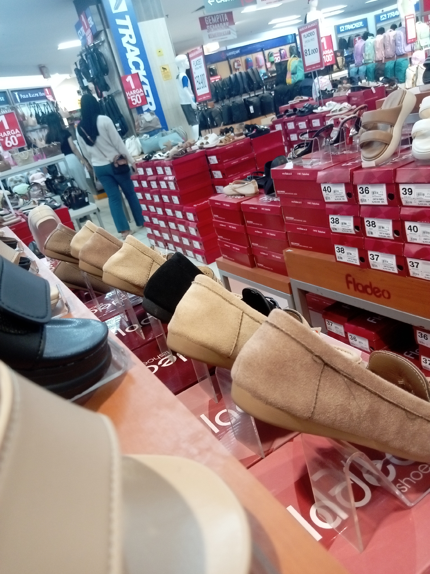 The image captures an indoor retail environment, likely a shoe store, located in Batam, Indonesia. In the foreground, several pairs of women's shoes are displayed on racks and in red boxes. These shoes appear to be loafers and sandals, with materials like suede and leather visible. Some shoes are presented on transparent stands.
Behind the footwear display, a woman wearing a white long-sleeved shirt, a black hijab, and blue jeans is walking away from the camera. She is carrying a brown handbag. Further in the background, there are aisles with more merchandise, including bags and other clothing items, displayed on shelves and hanging racks. Signage in Indonesian is visible, including "TRACKER," "MURAH BANGET," and prices like "81.000" and "100." There are also mannequins and stacks of red shoe boxes with labels indicating sizes and brands. The overall impression is of a busy retail setting in a department store or a specialized shoe shop.