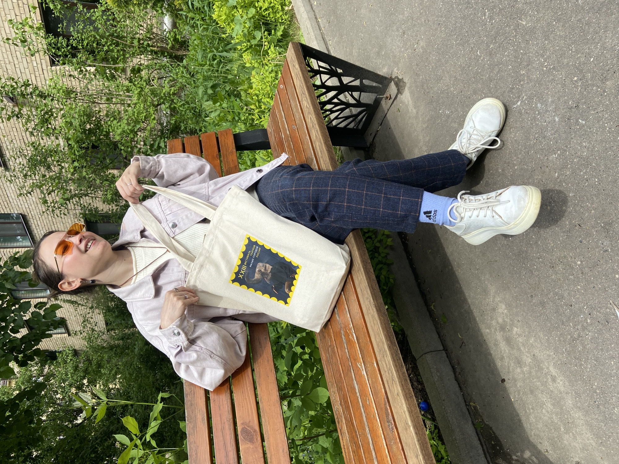 A person is lying on a wooden park bench in Peresvet, Russia, on a sunny day. The person is wearing a light purple jacket, a white tank top, blue plaid pants, light blue socks with an Adidas logo, and white sneakers. They are also wearing orange sunglasses. The person is holding a white canvas tote bag that has a yellow framed image with Russian text. The bench is located next to a sidewalk and a patch of green foliage. A light-colored brick building is visible in the background, partially obscured by trees. A black metal object is attached to the bench.