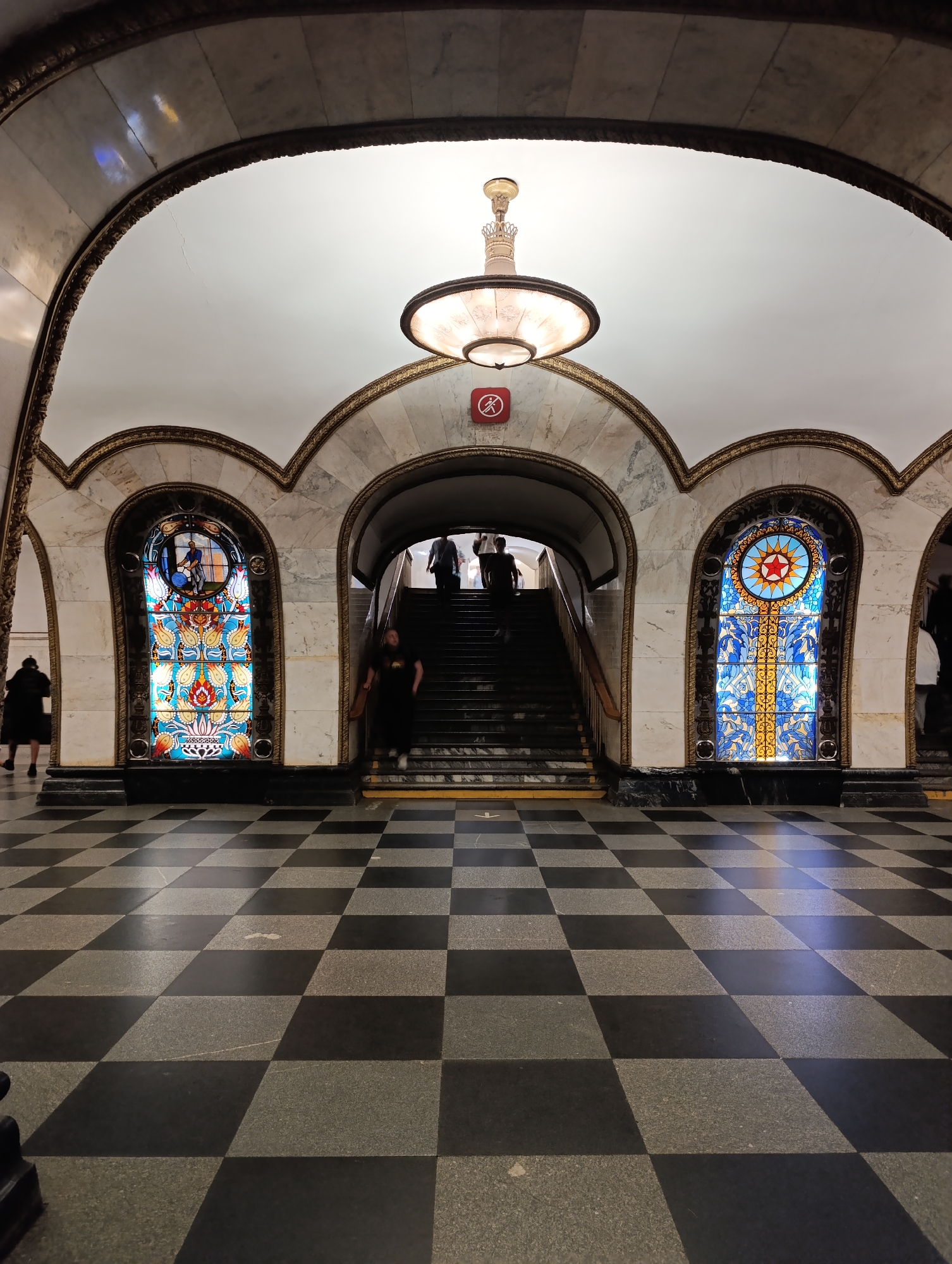 The image depicts an ornate underground passage or metro station, characterized by its classical architecture and opulent decorations, consistent with Moscow's historical metro system. The scene is dominated by a grand central staircase leading upwards, flanked by richly adorned walls and arches.
Several individuals are present, some ascending and others descending the dark-colored staircase. Their faces are indistinct, and their clothing is dark, suggesting a normal flow of pedestrian traffic.
The most striking features are the two large, arched stained-glass windows on either side of the staircase. The left window features a vibrant blue, orange, and red composition, with a lotus-like pattern at its base and a circular section at the top containing a stylized depiction of a worker. The right window, similarly arched, showcases a prominent red star within a circular motif, surrounded by intricate blue and yellow geometric and floral designs. These windows are set within marble walls and framed by elaborate gilded moldings.
Above the central staircase, a large, ornate chandelier with multiple tiers and glass panels hangs from the vaulted ceiling, casting a warm light. The ceiling itself is white and arched, with gilded trim tracing the contours of the arches. The walls are constructed from marble with visible veining.
The floor features a distinctive black and grey checkered pattern, contributing to the grand aesthetic of the space. Above the staircase entrance, a small red circular sign with a white border and a crossed-out figure is visible, likely indicating a prohibition such as "no loitering" or "no standing." The artificial lighting suggests it's an indoor or subterranean environment, with no discernible time of day or weather conditions.