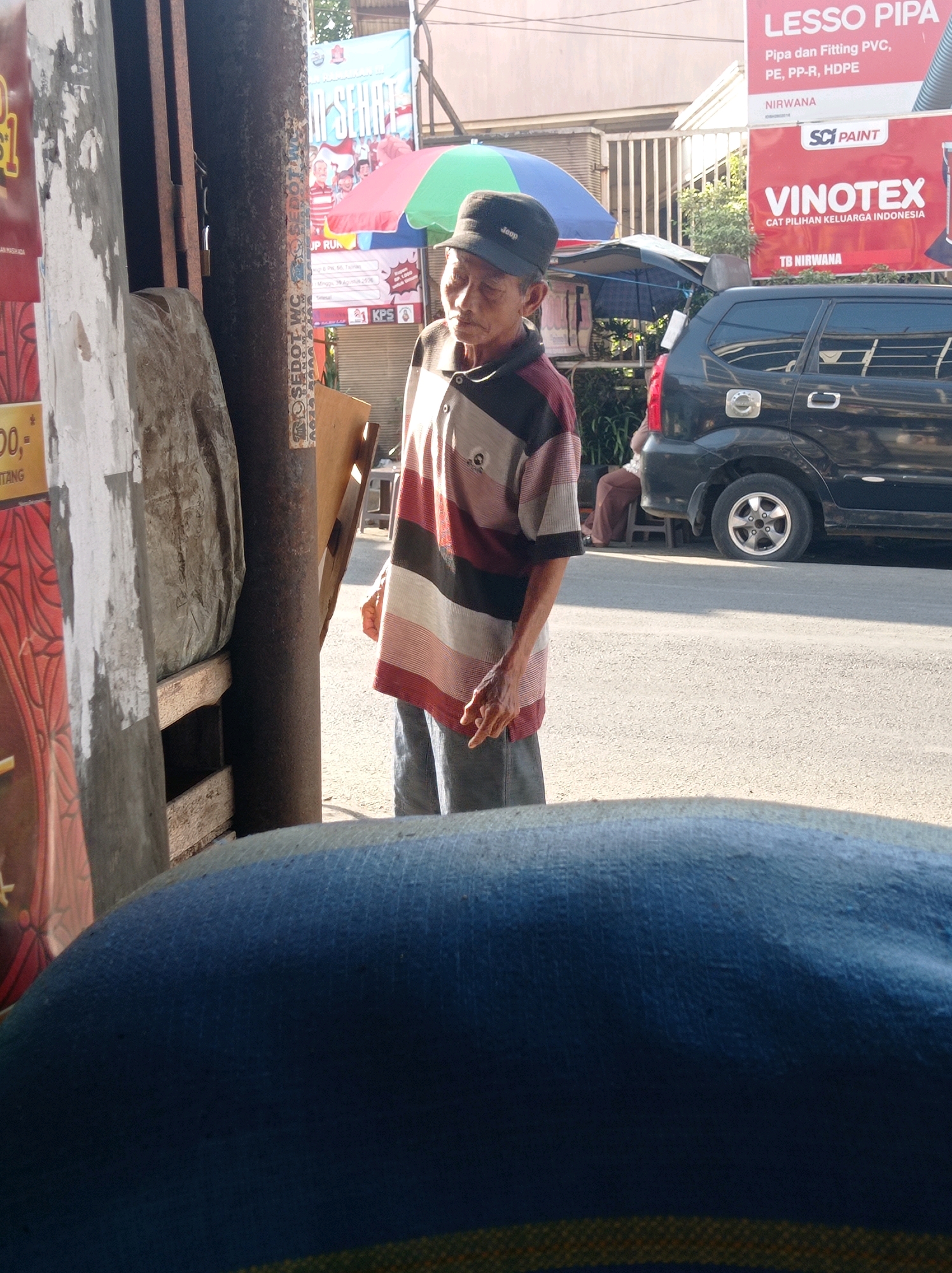 An elderly Asian man wearing a striped short-sleeved shirt, dark trousers, and a black baseball cap stands on a sidewalk next to a street in Tajinan, Indonesia. He is looking down and to his left, with his right hand near his chest. Behind him, a black SUV is parked on the street. In the background, a building with signage is visible. The overall scene appears to be an outdoor market or commercial area.