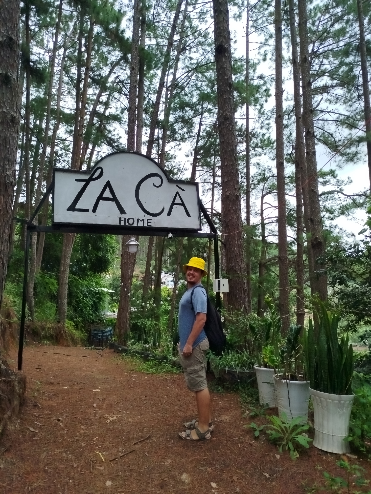 A man wearing a yellow bucket hat, a blue t-shirt, and khaki shorts stands on a dirt path in a pine forest. He is facing the camera and smiling, with a black backpack on his shoulder. Behind him, a sign reads "LA CÀ HOME". The path is lined with potted plants and trees. The scene appears to be a tourist attraction or a private residence in Da Lat, Vietnam, known for its pine forests and cooler climate. The lighting suggests it is daytime, possibly overcast.