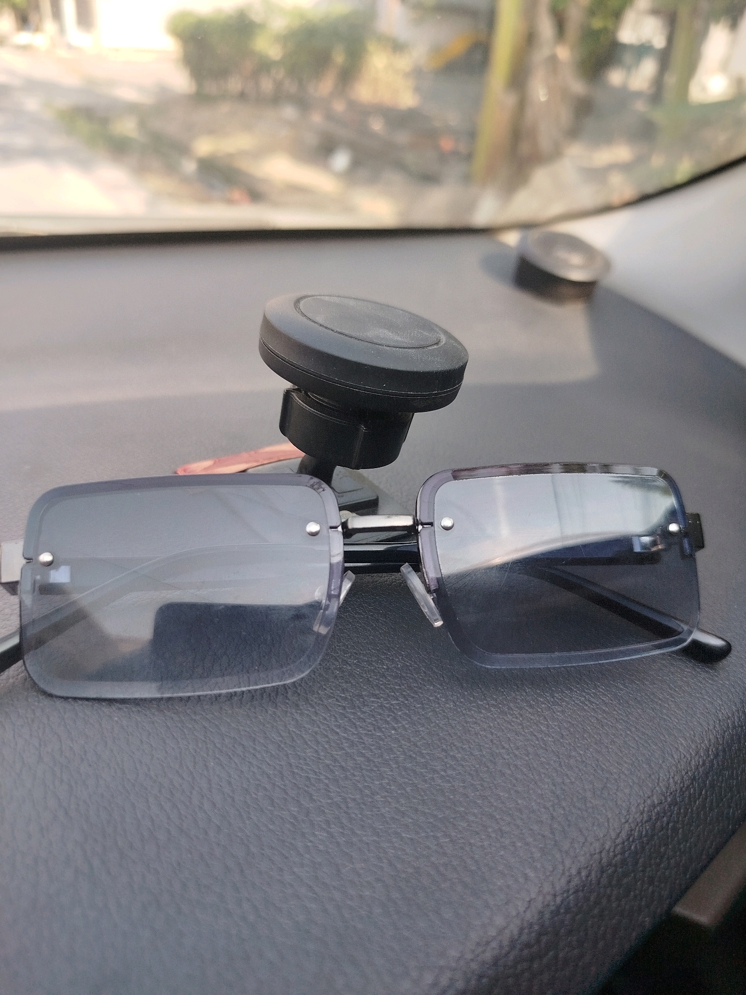 The image shows a pair of dark, rectangular sunglasses resting on a textured, dark gray dashboard. A black magnetic phone mount is positioned behind the sunglasses. The background is blurred, showing a view through a car windshield, with hints of foliage and a street or building in the distance. The lighting suggests an outdoor, daytime setting. The location is the city of Cagakagung, Indonesia.