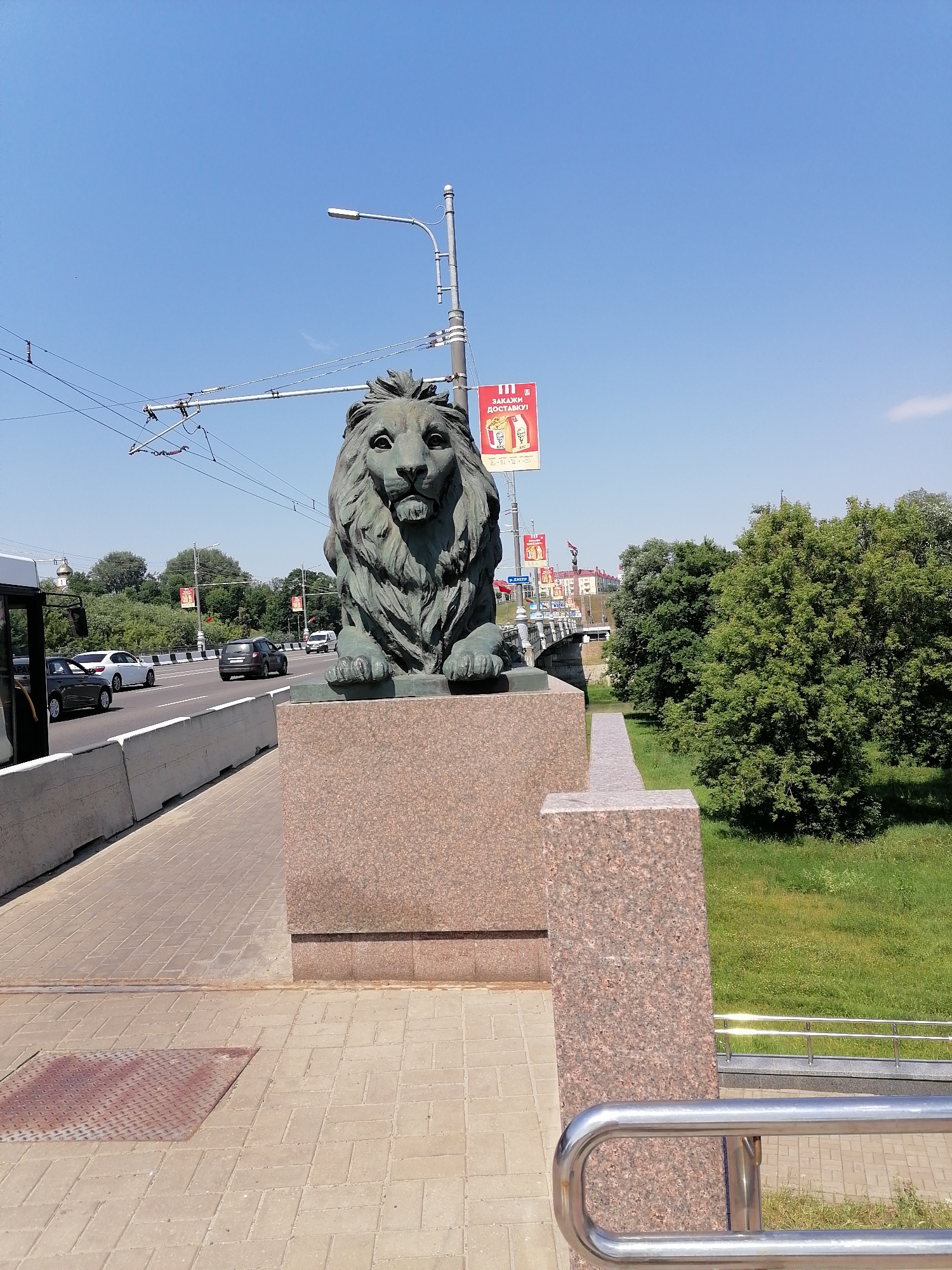 An outdoor, eye-level, medium shot captures a bronze lion statue situated on a granite pedestal. The statue is positioned on the right side of the frame, overlooking a street with vehicles. The lion faces forward, its mane detailed and its expression stern. Behind the statue, a street with multiple lanes of traffic is visible, with cars driving in both directions. A pedestrian walkway made of light-colored pavers runs alongside the road, bordered by a concrete barrier. In the background, lush green trees and buildings can be seen under a clear blue sky. A street lamp with overhead wires is also present. A banner advertising a fast-food chain is attached to a pole on the left side of the image. The overall scene depicts an urban environment with a prominent statue as a focal point.