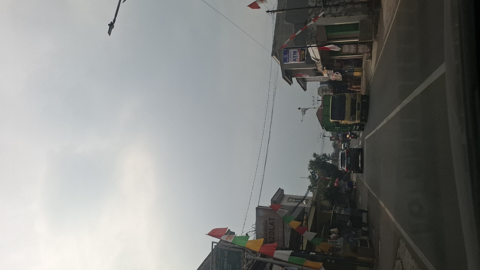 The image depicts a street scene in Sukasari, Indonesia, under an overcast sky. The viewpoint is from inside a vehicle, looking forward. The road is asphalt with white lane markings. On the left side of the road, there are buildings with storefronts adorned with colorful flags. One building displays a sign that reads "AZOLAT". Several small carts or stalls are present near the storefronts. Traffic on the road includes a black SUV in front, a yellow truck to the right, and several motorcycles. The buildings on the right side of the road also feature storefronts, with one displaying an ATM sign. Red and white Indonesian flags are visible along the street. The environment suggests a commercial or residential area within a town or city.