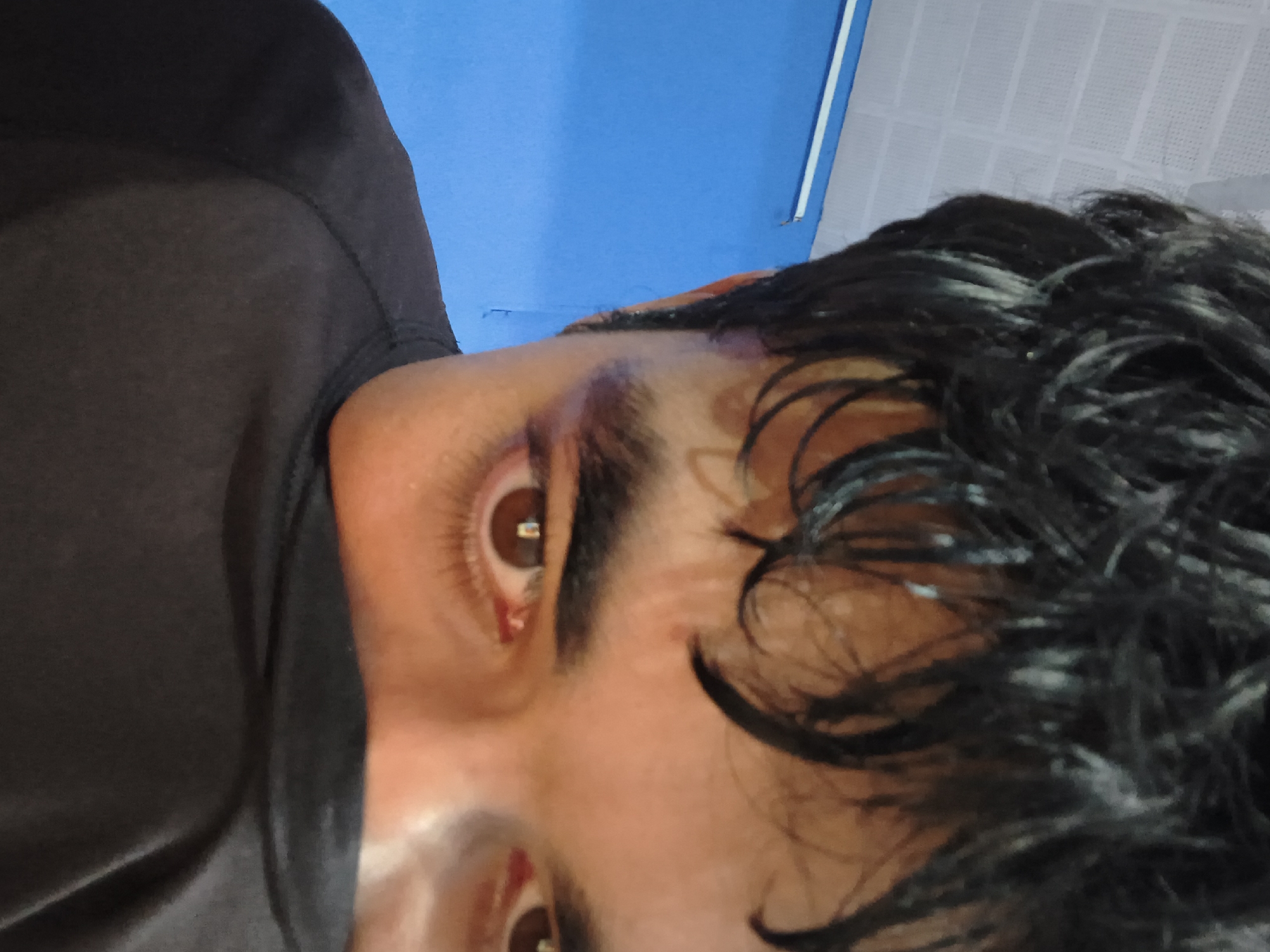 The image is a close-up, low-angle shot of a person's face, partially obscured by a black garment. The person has dark, messy hair and brown eyes. The background consists of a bright blue wall and a white ceiling with visible grid lines. The lighting suggests an indoor environment. The person is wearing a black t-shirt. No specific location in Dharamshala, India is discernible from the image.
