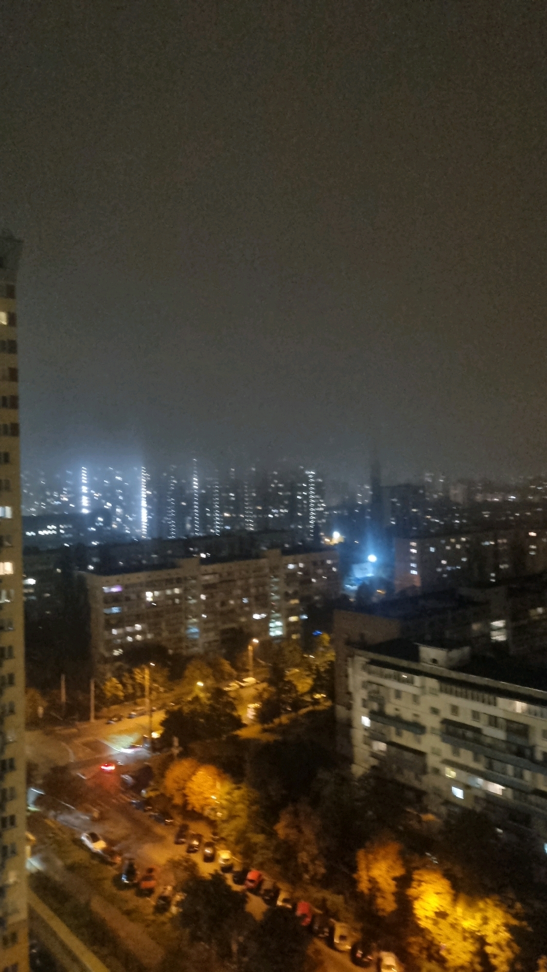 This is an aerial, nighttime photograph of a city skyline, likely in Kyiv, Ukraine. The image is taken from a high vantage point, looking down and across a densely populated urban area. The sky is overcast and dark, with a hazy or foggy quality that softens the lights of the distant buildings.
In the foreground and mid-ground, numerous apartment buildings are visible. These buildings are multi-story structures with many windows, most of which are illuminated, indicating that they are occupied. The architecture appears to be typical of Soviet-era apartment blocks, with repeating facades and balconies. Some buildings are closer and more detailed, while others recede into the background, becoming less distinct.
Below the apartment buildings, there are streets with parked cars and moving vehicles. Streetlights illuminate the roads and sidewalks. There are also trees and other foliage scattered throughout the urban landscape, some of which are illuminated by artificial lights, giving them a yellow or orange hue.
In the distance, a large cluster of taller buildings is visible. These buildings are lit up with numerous lights, creating a shimmering effect against the hazy sky. The density and height of these structures suggest a commercial or more modern residential area. A bright, bluish light emanates from somewhere within this distant cluster, possibly from a sports facility, a large event, or industrial lighting.
The overall impression is one of a bustling city at night, with a considerable amount of artificial light contributing to its illumination. The weather appears to be damp or foggy, as suggested by the diffused light and the softened appearance of distant objects. There are no visible people in the image.