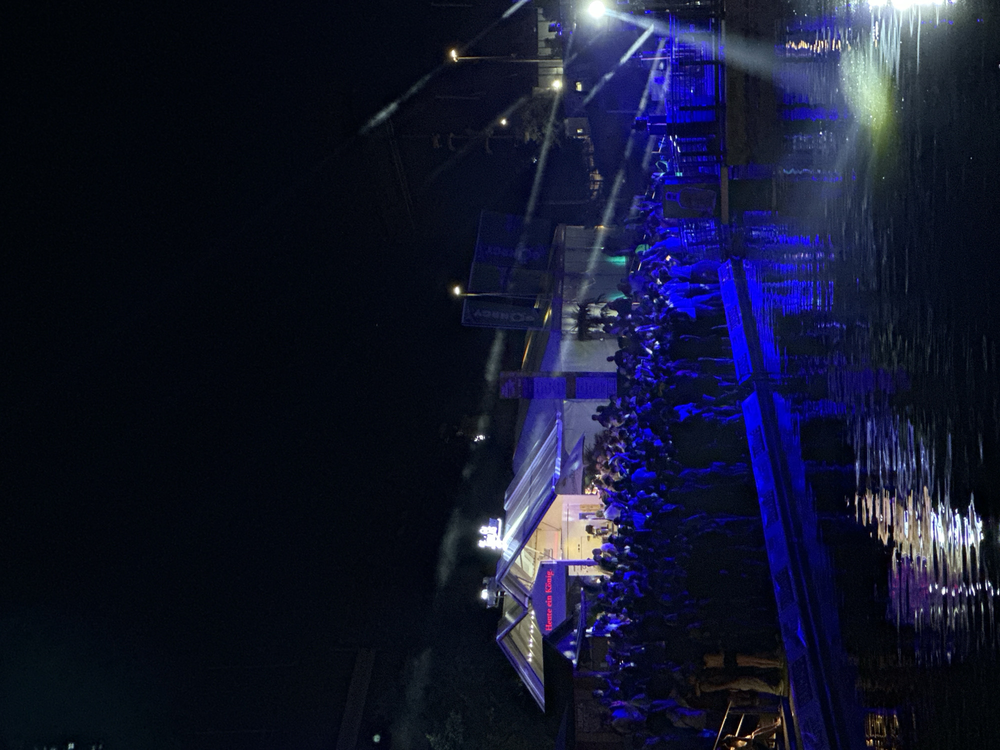 A nighttime scene depicts an outdoor event, likely a concert or festival, taking place near a body of water. The foreground features the dark, rippling surface of the water, reflecting the blue and white lights from the event. A low, dark blue structure, possibly a stage or platform, runs along the edge of the water. In the midground, a large crowd of people, illuminated by the event's lighting, are gathered in front of a building with a transparent, multi-pitched roof. Red neon lettering on the building reads "Heute ein König." Further back, more structures and lighting equipment are visible, along with trees and the dark silhouette of a bridge or elevated structure. The overall atmosphere is that of a vibrant, illuminated nighttime gathering. This scene is consistent with events held at the "Pumpe" cultural center in Kiel, Germany, which is located near the Kiel Fjord.
