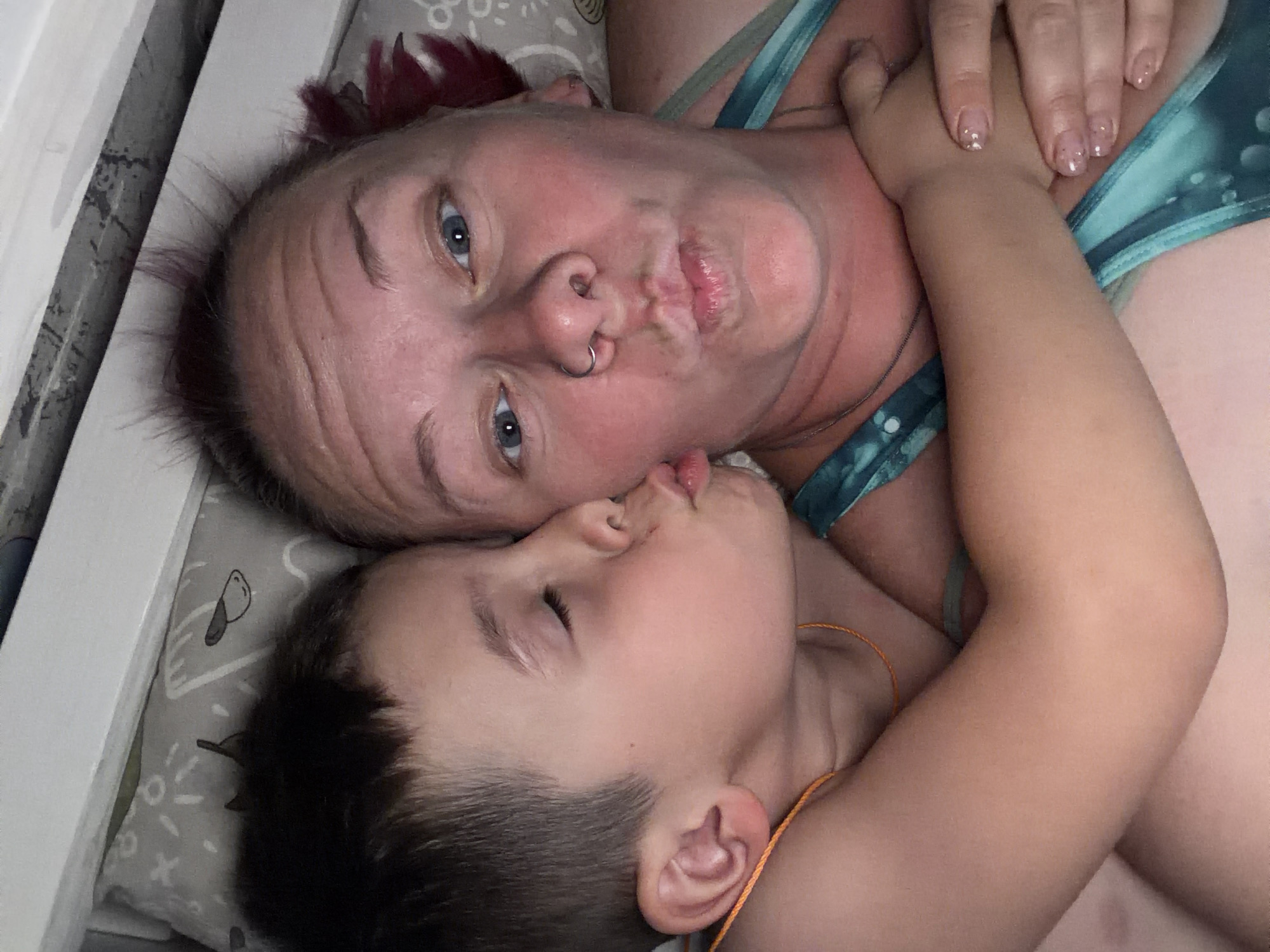A woman with fair skin and red-tinged hair is lying on a patterned pillow next to a young boy. The woman has blue eyes, a nose ring, and is puckering her lips. The boy has short, dark hair and closed eyes, and he is lying with his head near the woman's face. The woman is wearing a blue and green patterned top. The location is not discernible from the image.