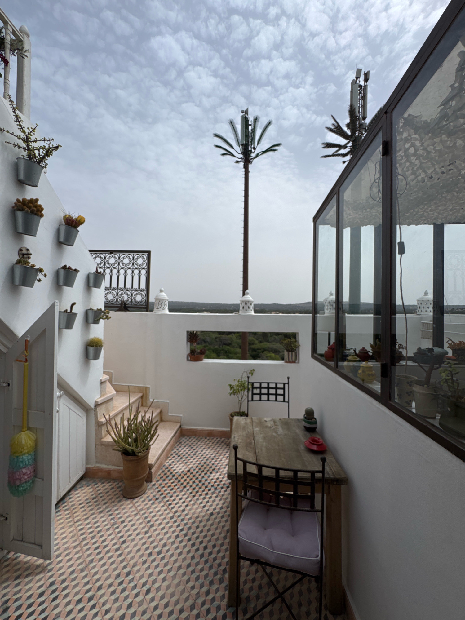 The image depicts an outdoor terrace or balcony area with a view of the sky and a distant landscape. The terrace features white walls, a tiled floor with a geometric pattern in shades of pink, grey, and white, and a set of stairs leading up. Several potted plants, including succulents and cacti, are arranged on the wall and near the stairs. A wooden table with chairs is set up on the terrace, and a glass-enclosed area with more plants and decorative items is visible. In the background, two telecommunication towers with antenna arrays are prominent against the cloudy sky, with palm trees nearby. The architecture suggests a Moroccan style, consistent with the location in Essaouira. The scene is static, with no people or animals present. The overall impression is a well-decorated outdoor living space with a functional yet scenic setting.