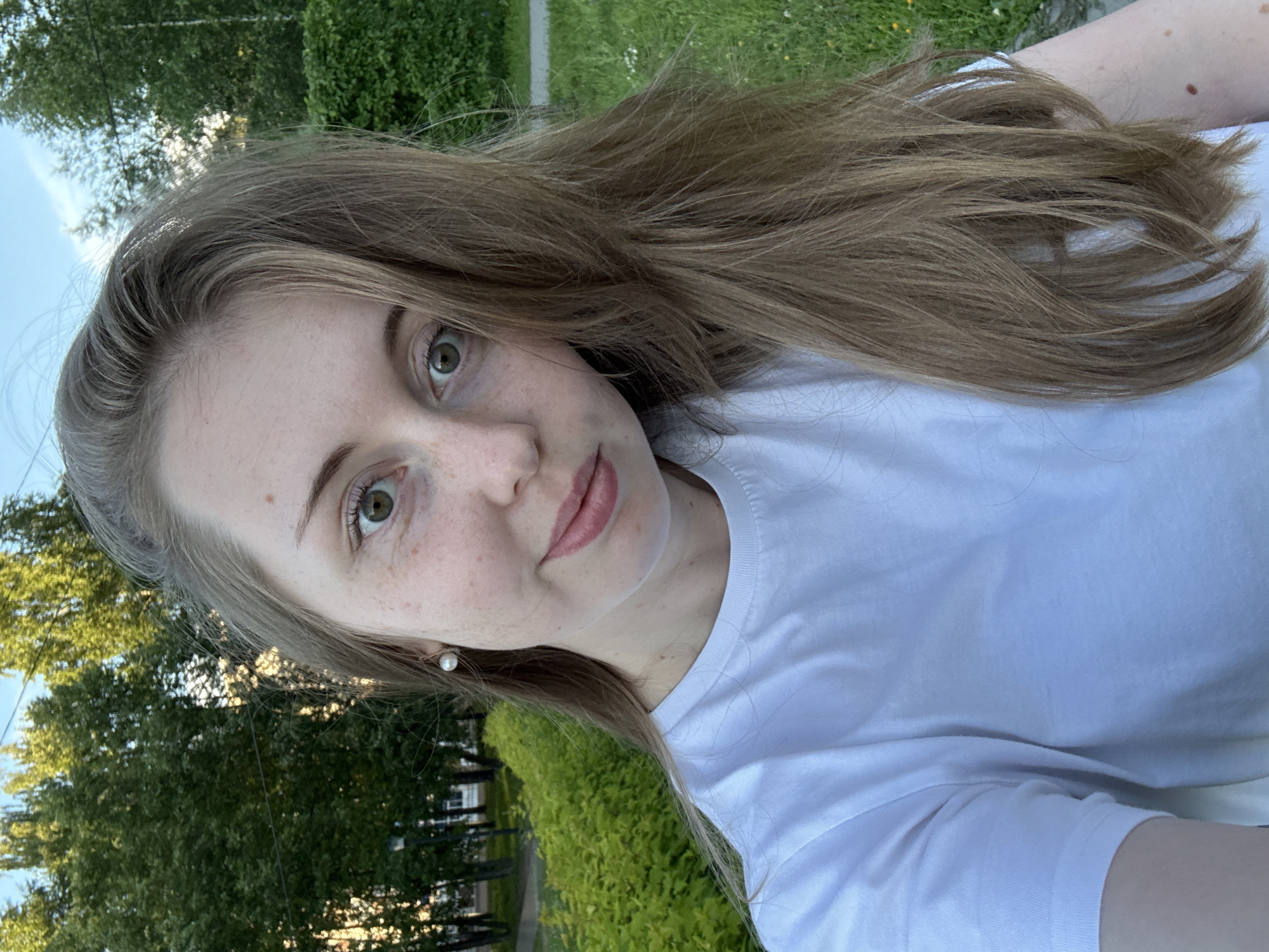 An outdoor, medium shot captures a young woman with light brown hair and green eyes posing for a selfie in a park-like setting. She is wearing a white t-shirt and a pearl earring on her left ear. Her face is turned slightly towards the camera, with a subtle smile. The background consists of lush green trees and foliage, with a clear blue sky visible at the top. The lighting suggests it is either late afternoon or early evening.