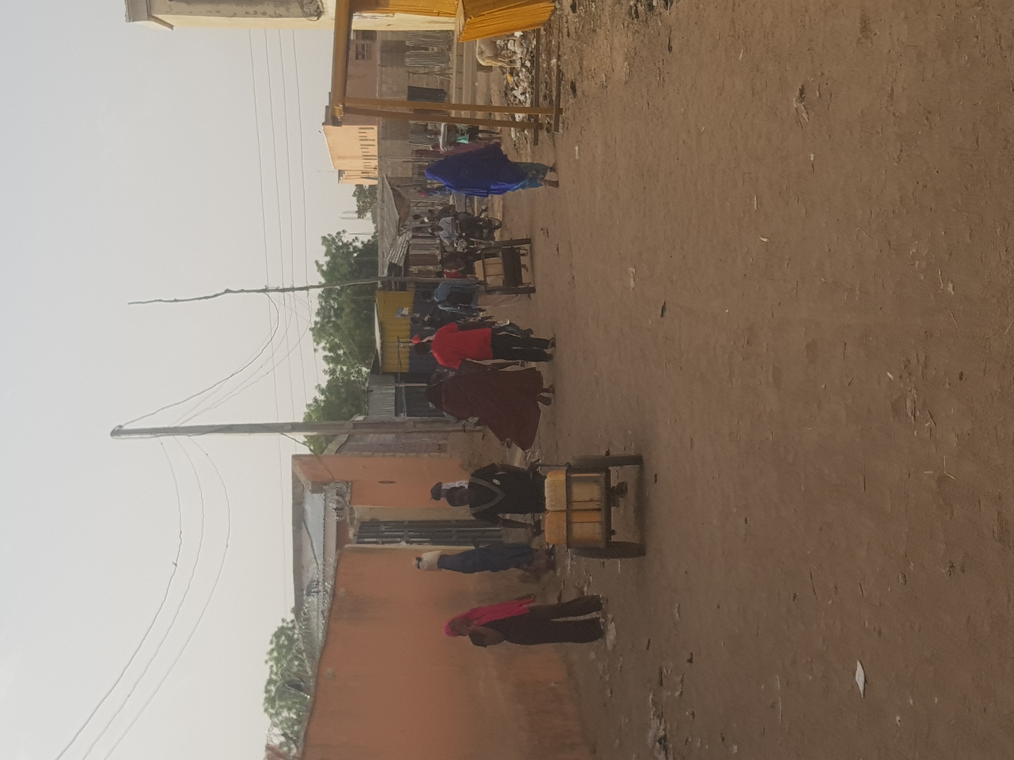 The image depicts a street in Dabawa, Nigeria. The ground is dirt, and there are buildings on either side of the street, some of which appear to be made of mud or other simple materials. A utility pole is visible with overhead wires running along the street. Several people are present, some wearing colorful clothing, including a woman in a blue robe. There are individuals moving along the street, and one person is pushing a cart with yellow containers. Some other people are sitting along the side of the road as well. There is trash visible in the street and around the buildings. In the distance, there appear to be more buildings and trees. The sky is overcast, suggesting the possibility of dry or dusty conditions.