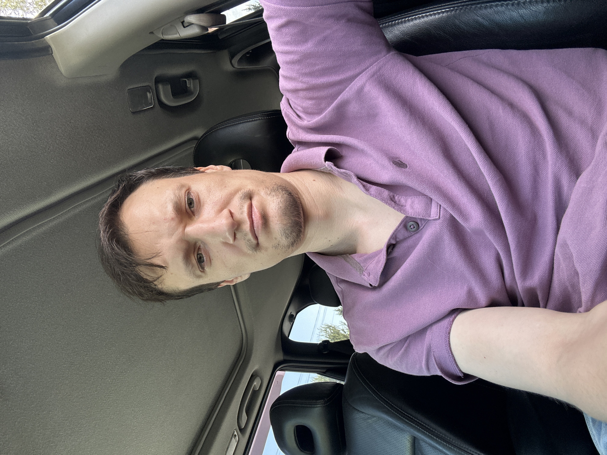 A man in a purple polo shirt sits in the driver's seat of a car, looking towards the camera. The car's sunroof is open, and the interior is visible, including black leather seats and the gray headliner. The scene appears to be outdoors, with trees and a bright sky visible through the windows. The overall impression is of a casual selfie taken inside a vehicle. The location is the city of Makiv, Ukraine.