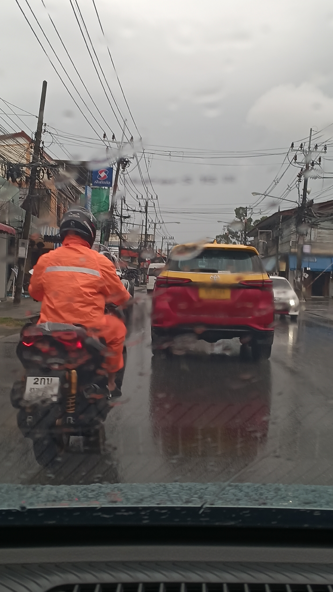 The image captures a street scene in Ko Samui District, Thailand, during a rainy day. The foreground shows a view through a wet windshield, with raindrops visible. A person on a scooter, wearing an orange rain suit and a black helmet, is prominently featured in the middle ground, moving away from the viewer. The scooter has a license plate with Thai characters and the number "2vu". To the right of the scooter, a red and yellow taxi is traveling in the same direction. In the background, there are buildings, utility poles with numerous wires, and other vehicles on the wet road. The sky is overcast, indicating cloudy weather. The scene is a typical urban street with traffic, infrastructure, and signs of recent rainfall.