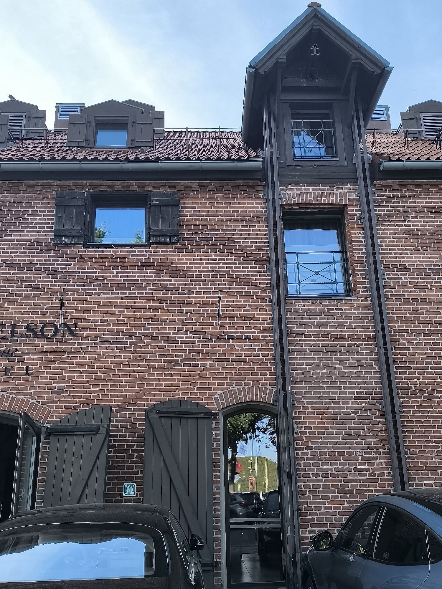 The exterior of a brick building with dark wooden shutters and windows is shown, with two cars parked in front. The building appears to be a historical structure, possibly a former industrial building, with a mansard roof and dormer windows. The windows reflect the blue sky and trees. On the left side of the building, a sign with the letters "ELSON" is visible. A green exit sign is mounted on the wall near a doorway. The sky is overcast. The location is identified as Klaipėda, Lithuania.