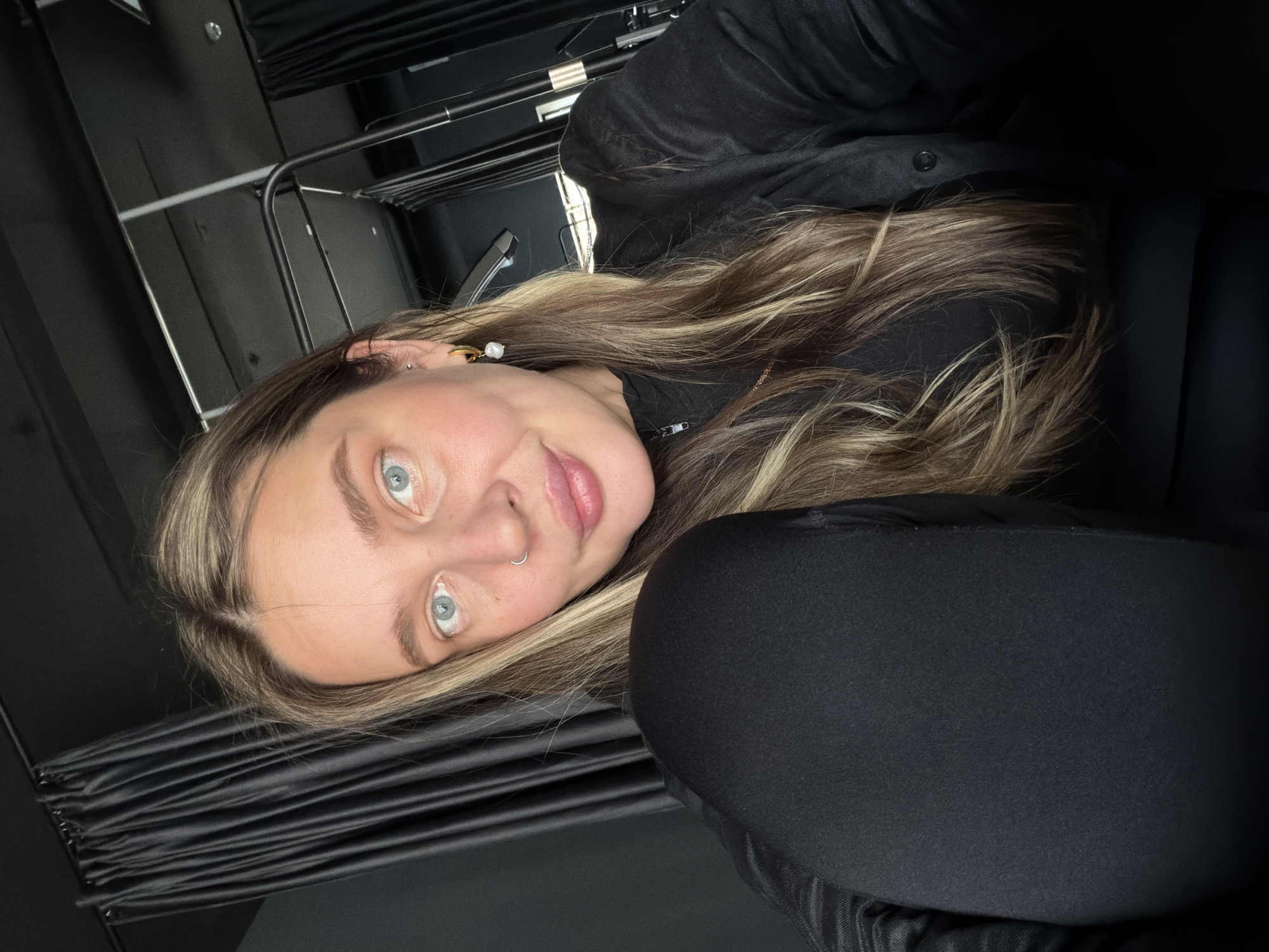 A selfie of a fair-skinned woman with light blue eyes and blonde-highlighted hair, is taken in what appears to be the backstage area of a theater. She is wearing a black jacket and possibly black leggings or pants. A silver nose ring is visible in her left nostril, and a gold earring with a pearl accent is visible in her left ear. The background reveals black curtains and metal structures, typical of theater set construction. There are no clear indications of a specific location in Moscow.