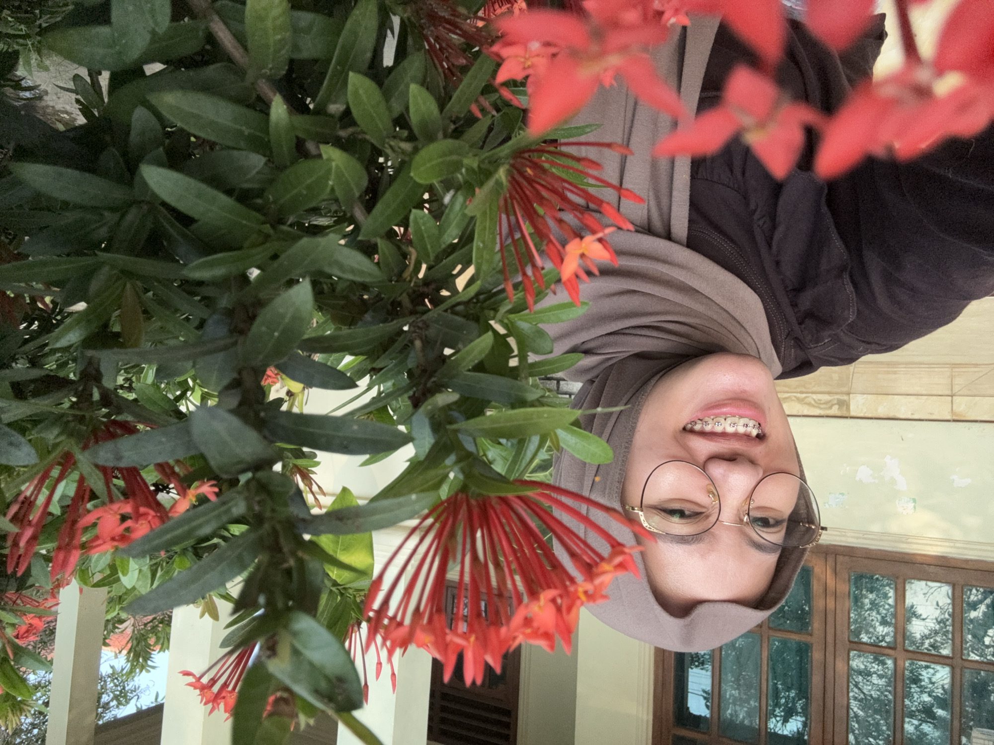 A young woman wearing a hijab and glasses smiles at the camera, her braces visible. She is wearing a dark brown top with ruffled details and a light brown hijab. The background features a window with multiple panes and a light-colored wall. To the right, a flowering plant with red blossoms and green leaves is in focus. The setting appears to be an outdoor area near a building.
