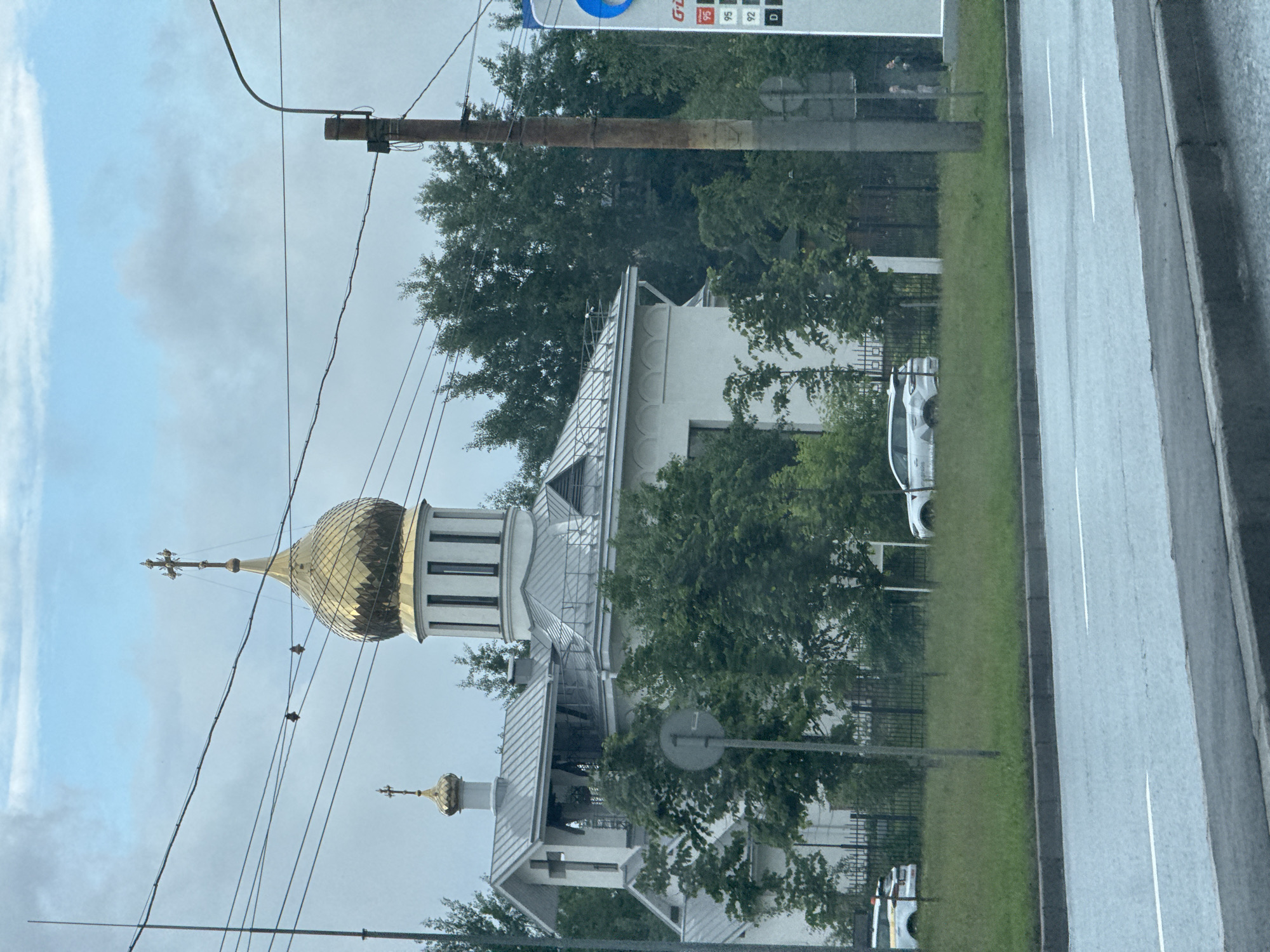A white church with a golden dome and a cross on top is visible behind a fence and trees. A street with cars in the foreground indicates an urban setting. The sky is overcast with some clouds. A utility pole with wires is also present in the scene. The image was taken in Saint Petersburg, Russia.