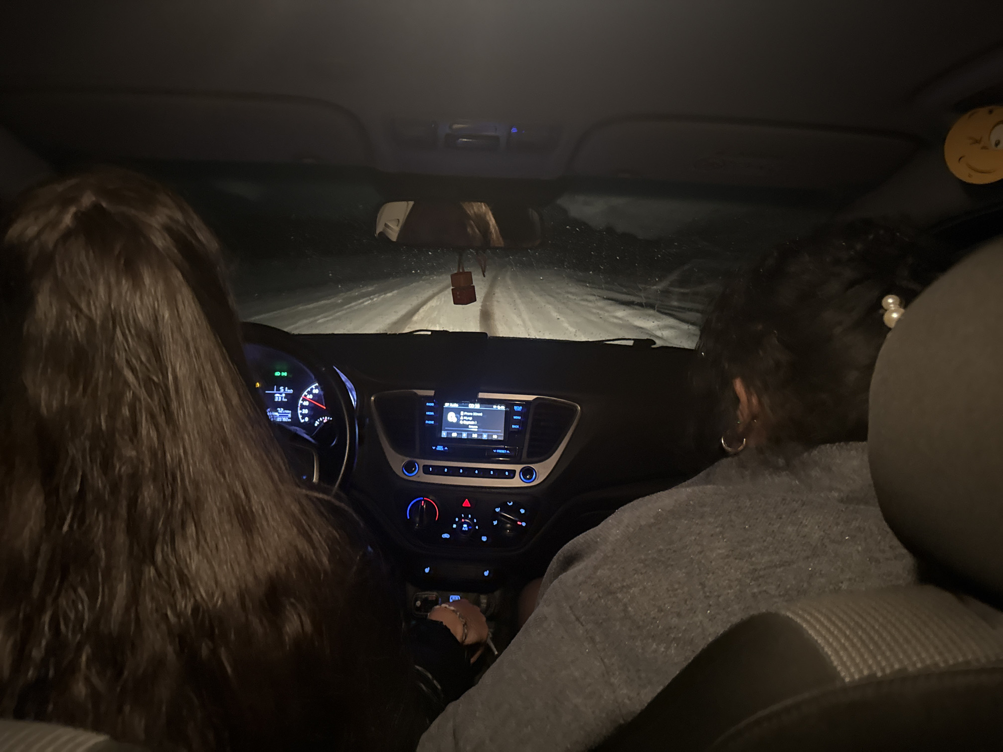 Two individuals are inside a car traveling on a snow-covered road at night. The dashboard is visible, showing the car's infotainment system and controls. The road appears to be in an area near Кемуль, Russia.