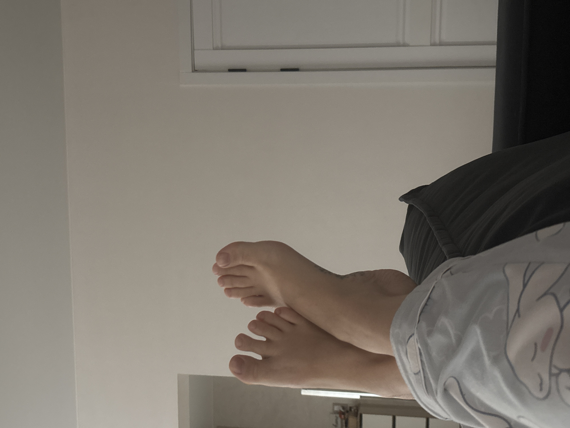 The image shows a pair of human feet resting on a dark gray cushion. The feet are pale in complexion, with visible toes. The individual is wearing blue and white patterned pajama pants. In the background, a white wall with a white window frame is visible. A portion of what appears to be a kitchen counter or other cabinetry can be seen in the lower part of the image. The image is rotated 90 degrees counterclockwise. The location is unknown, including whether it is in Chaikovsky, Russia.
