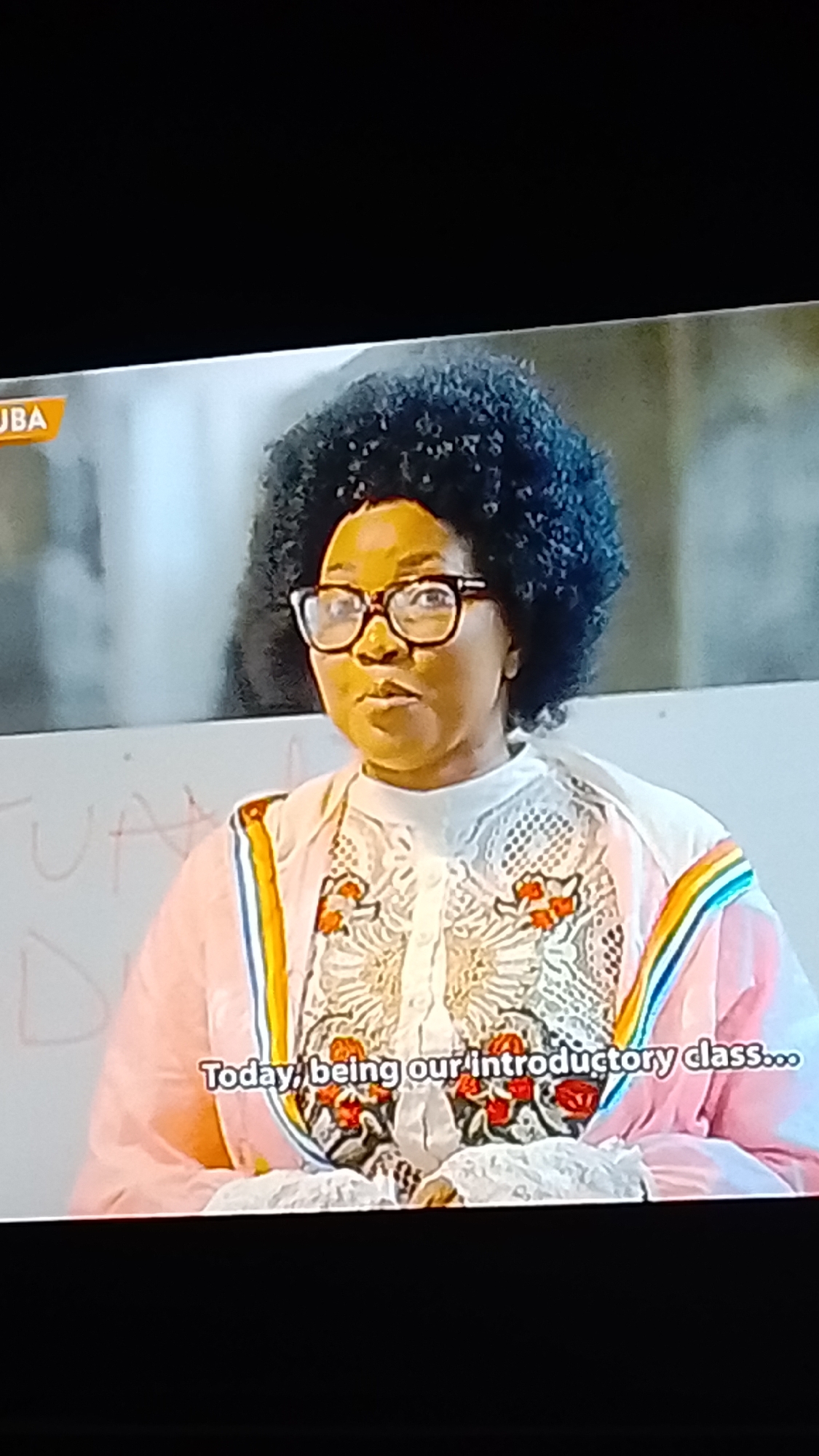 A medium shot captures a Black woman with a voluminous afro, wearing glasses and a pink and white patterned shirt. She appears to be speaking to an audience. In the background, a whiteboard with faint writing is visible. Text overlays the bottom of the image, reading "Today, being our introductory class...". The scene takes place indoors, potentially a classroom or lecture hall. There is no information to suggest the location is in Lokoja, Nigeria.