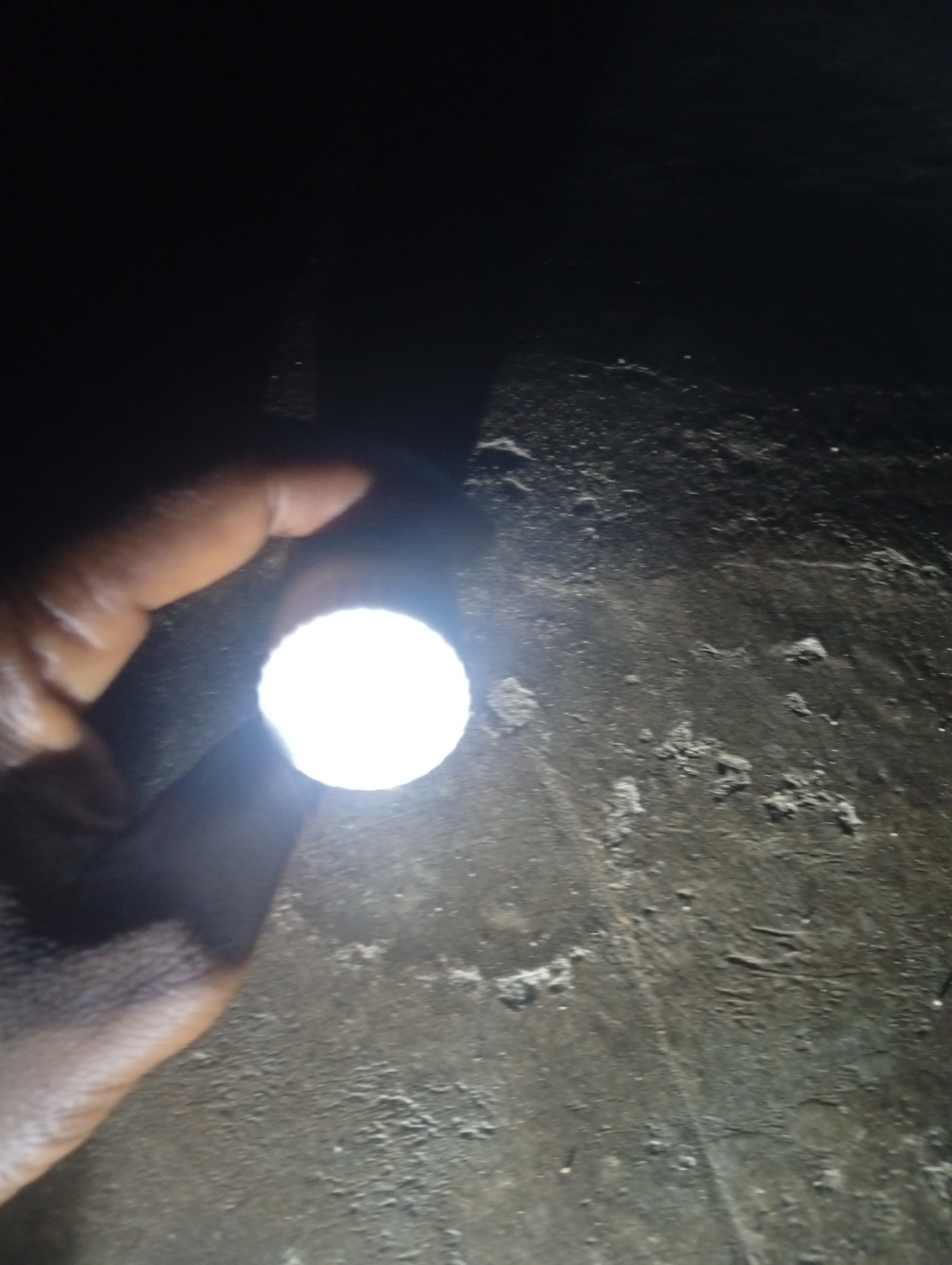 In Jalingo, Nigeria, a close-up shot shows a hand holding a bright, circular object that is emitting light. The object is likely a flashlight or the lens of a light source. The hand, with dark skin, is positioned in the lower left quadrant of the image, partially obscuring the light source. The background is dark and textured, appearing to be a rough, uneven surface, possibly a wall or ground. The lighting from the object casts a strong illumination on the immediate surroundings, creating a stark contrast between the bright light and the dark environment. No other individuals are visible in the frame.
