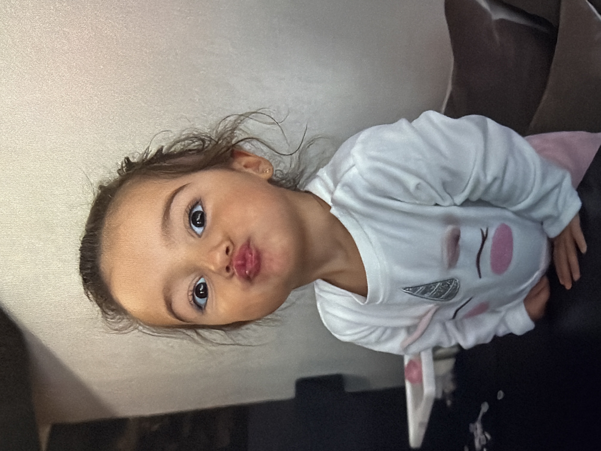 A young child with brown hair and eyes is puckering their lips in a kissing gesture. The child is wearing a white long-sleeved shirt with a unicorn design on the front. The child is sitting or lying on a dark-colored surface, possibly a couch or bed, next to a light-colored wall. The location is Unknown, Unknown.