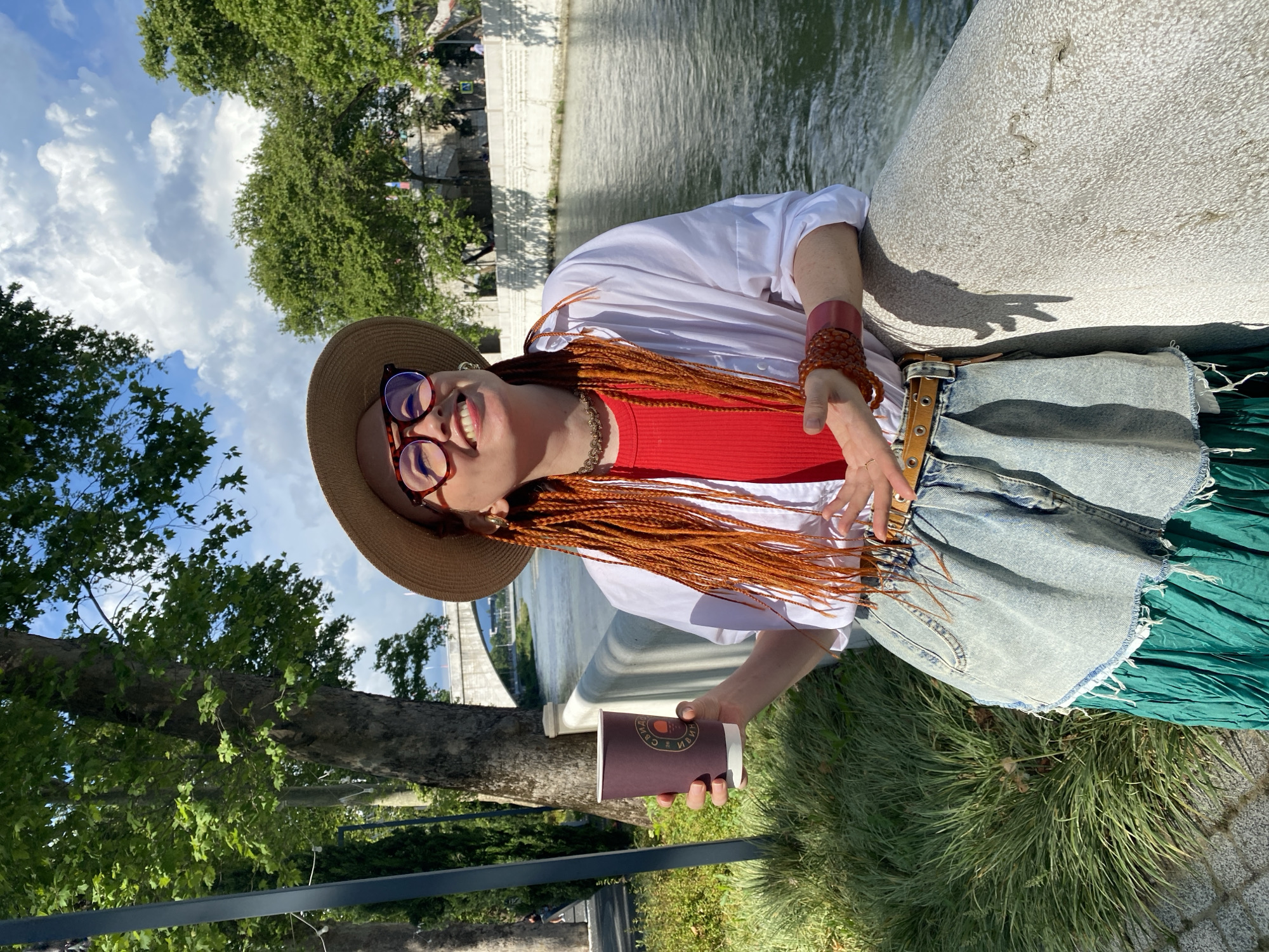A woman with bright orange braided hair, wearing a straw hat, red glasses, a red top, a white unbuttoned shirt, a denim skirt, and a green skirt, stands by a river in Sochi, Russia. She holds a brown paper cup in her left hand and leans on a stone railing. The background shows a river, trees, and a bridge. The sky is partly cloudy.