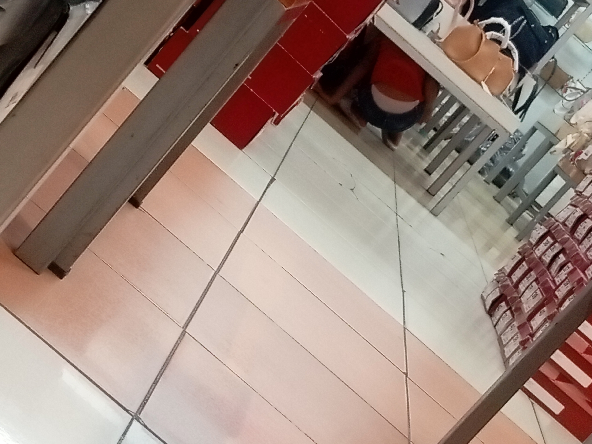 The image shows a retail environment, likely a department store or boutique, with displays of merchandise. The floor is tiled in a light color, possibly beige or cream, with darker grout lines forming a grid pattern. In the foreground, there are two slanted gray metal shelves. On the shelves, several items are visible, including a light brown handbag and several dark blue bags. A person is partially visible, wearing a red top and dark shorts. They appear to be in a fitting room or browsing area. In the background, there are red display shelves and stacks of boxed merchandise, some with white labels. The overall lighting suggests an indoor setting. The location is identified as Batam, Indonesia.