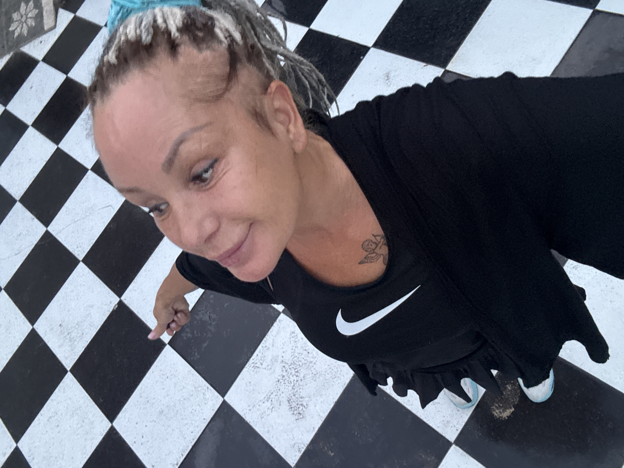 A woman with gray dreadlocks tied up with a teal band is standing on a black and white checkered floor, possibly in Kuta, Indonesia. She is wearing a black Nike sports bra with a small angel tattoo on her chest, and a black open-front cardigan or jacket. Her right hand, with pink painted nails, is extended towards the floor, and her thumb is extended as if giving a thumbs-up. She is wearing black pants and white sneakers with light blue accents. The woman is looking upwards and to her right, with a slight smile on her face. The checkered floor appears to be made of large tiles.