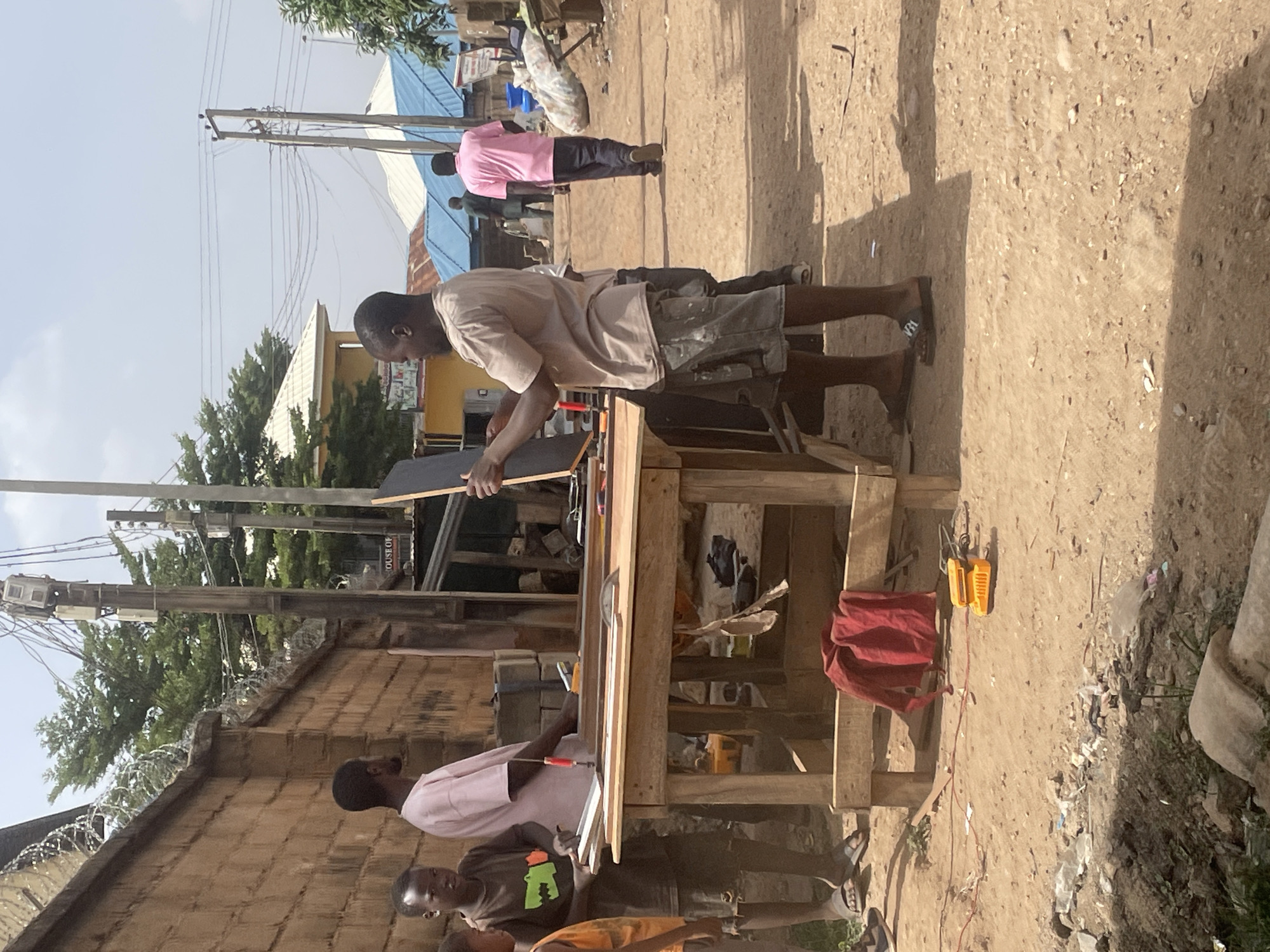 The image shows a scene in Jos, Nigeria, featuring several people gathered around a makeshift wooden table. A man is actively working on a black object on the table, possibly measuring or cutting it. Another man, in a pink shirt, is holding a similar black object. A young boy stands beside him, wearing a shirt with a green graphic. The table is surrounded by tools and objects, including a red cloth and a yellow electronic device with a wire on the ground. The ground is dirt, littered with debris. In the background, there are more people, buildings with corrugated metal roofs, trees, and electrical poles. A brick wall topped with barbed wire is visible on the left side of the image.
