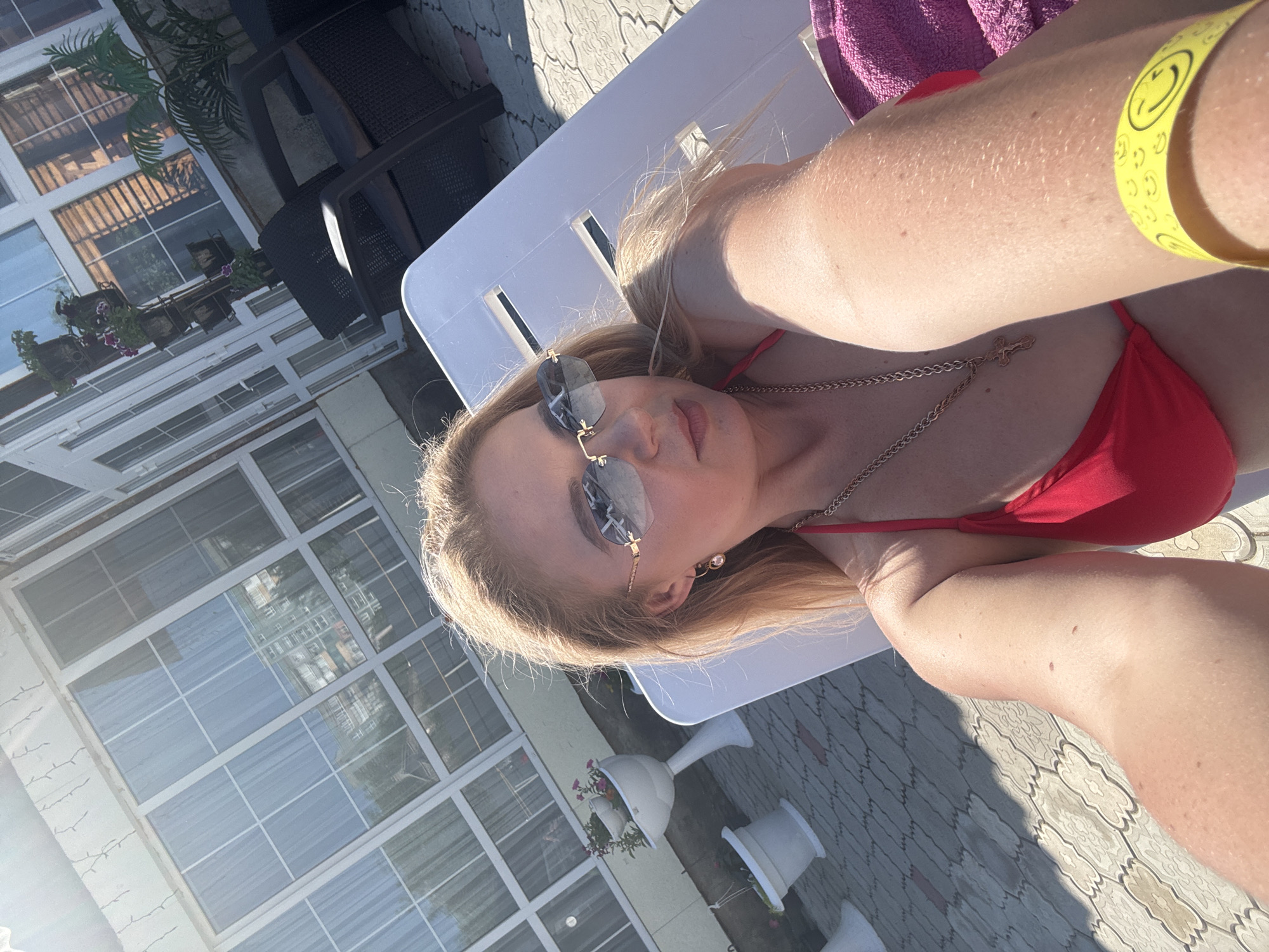 An outdoor selfie captures a blonde woman with fair skin and light makeup, wearing a red bikini top and gold-toned sunglasses with a hexagonal shape. She has a gold chain with a cross pendant around her neck and a pink towel is visible on her arm. A yellow wristband with smiley faces is on her right wrist. The woman is reclining on a white lounge chair. The background shows a tiled patio, a building with large windows, and potted plants. The lighting suggests it is daytime and sunny.