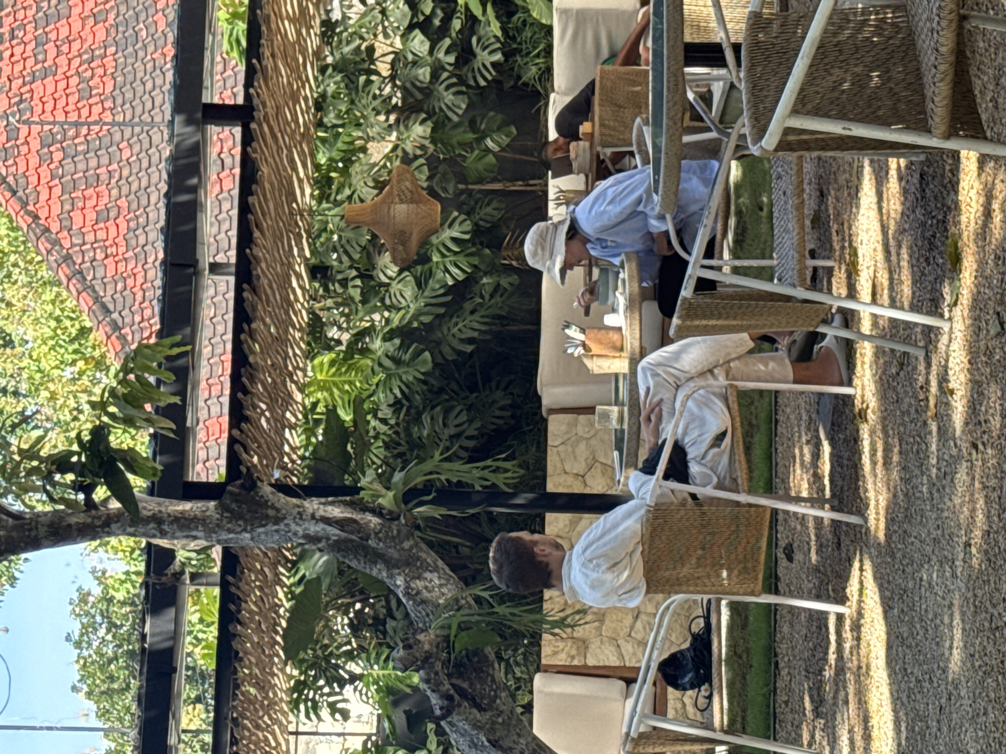 The image depicts an outdoor dining scene in Kuta Selatan, Indonesia. Two individuals, possibly a couple, are seated at a table. One person wears a light blue shirt and a sunhat, appearing to eat. The other, wearing a white shirt and pants, sits across. Wicker chairs surround the table, and the ground is covered in a textured, possibly gravel-like material. Lush tropical plants and a decorative woven lampshade adorn the space. Overhead, a red-tiled roof structure is visible. A tree trunk with foliage is to the left, adding to the outdoor ambiance.