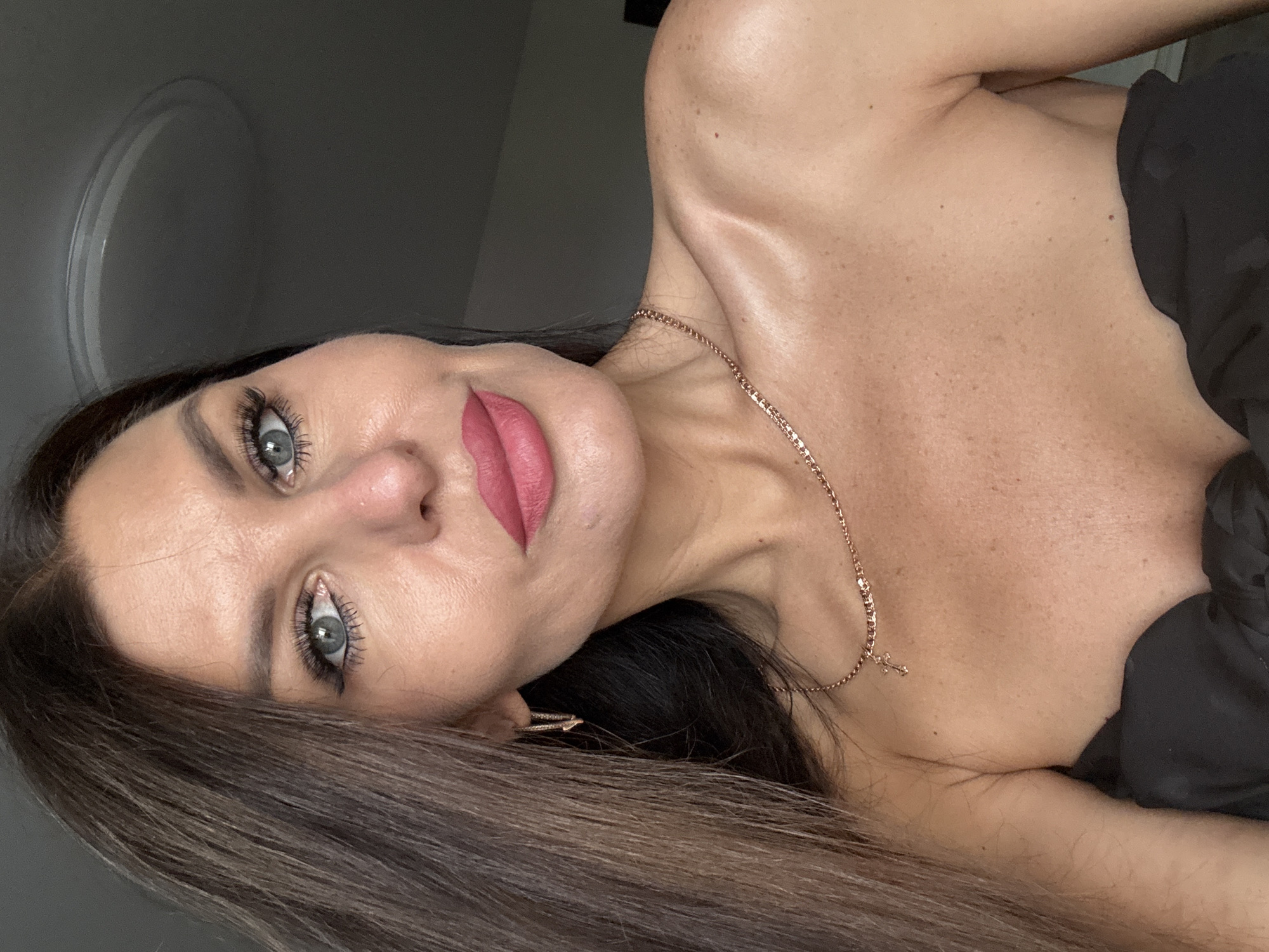This is a close-up, self-taken portrait of a woman with long, brown hair and light blue eyes. She is wearing pink lipstick and has a subtle smile. She is also wearing a thin gold necklace with a cross pendant and gold hoop earrings. The background is a plain gray wall with a circular light fixture. The image does not contain any information about the location in the city of Chaikovsky, Russia.