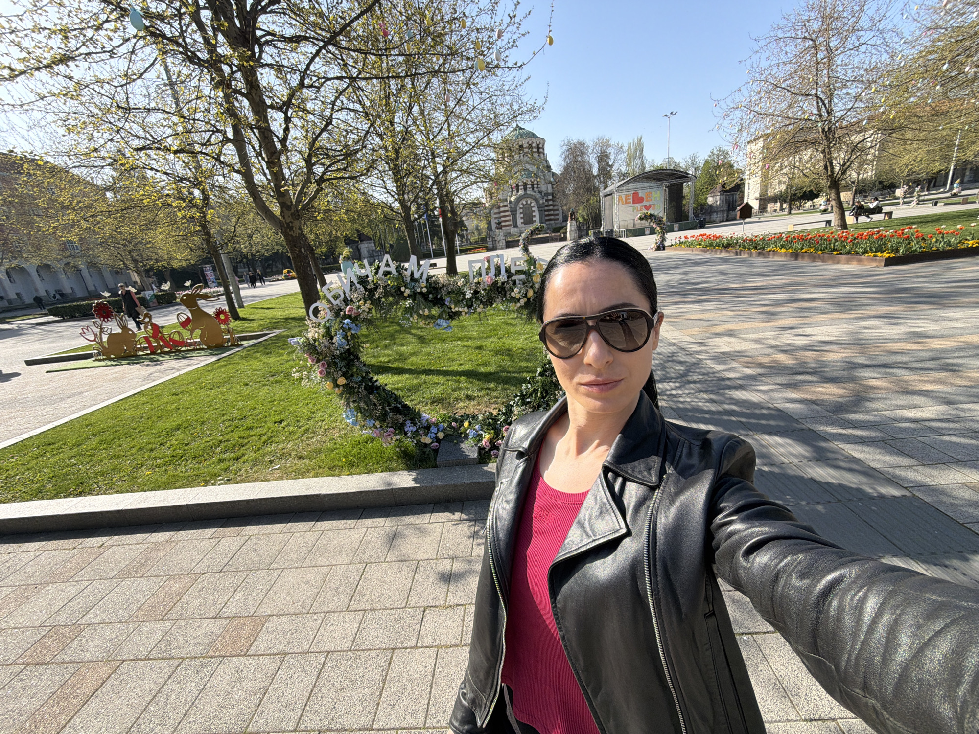 A woman is captured in a selfie in a sunlit public square in Pleven, Bulgaria. She is wearing sunglasses, a black leather jacket over a pink top, and has dark hair pulled back. Her expression is neutral.
The background reveals a lively park setting under a clear blue sky. A large, heart-shaped arch made of pastel flowers and greenery stands on the grass behind her. To the left of this arch, wooden cutouts of rabbits and flowers suggest an Easter or spring theme. Several trees, some with new green leaves and others still bare, are adorned with colorful egg decorations.
In the mid-ground, the distinctive architecture of the Mausoleum-Chapel "St. George the Conqueror" is visible with its domes and striped masonry. Further back, a stage is set up, displaying the word "ПЛЕВЕН" (Pleven) alongside a heart icon, and likely "ЛЮБИМ" (Beloved) as part of a larger message "ЛЮБИМ ПЛЕВЕН". The floral arch also features text, partially visible as "ОБИЧАМЕ ПЛЕВЕН" (We love Pleven).
Bright sunlight casts strong shadows across the paved walkways and grassy areas, indicating it is likely midday or early afternoon. Beds of orange and yellow tulips add vibrant color to the scene. Several people can be seen strolling in the background, enjoying the pleasant weather and festive decorations.
