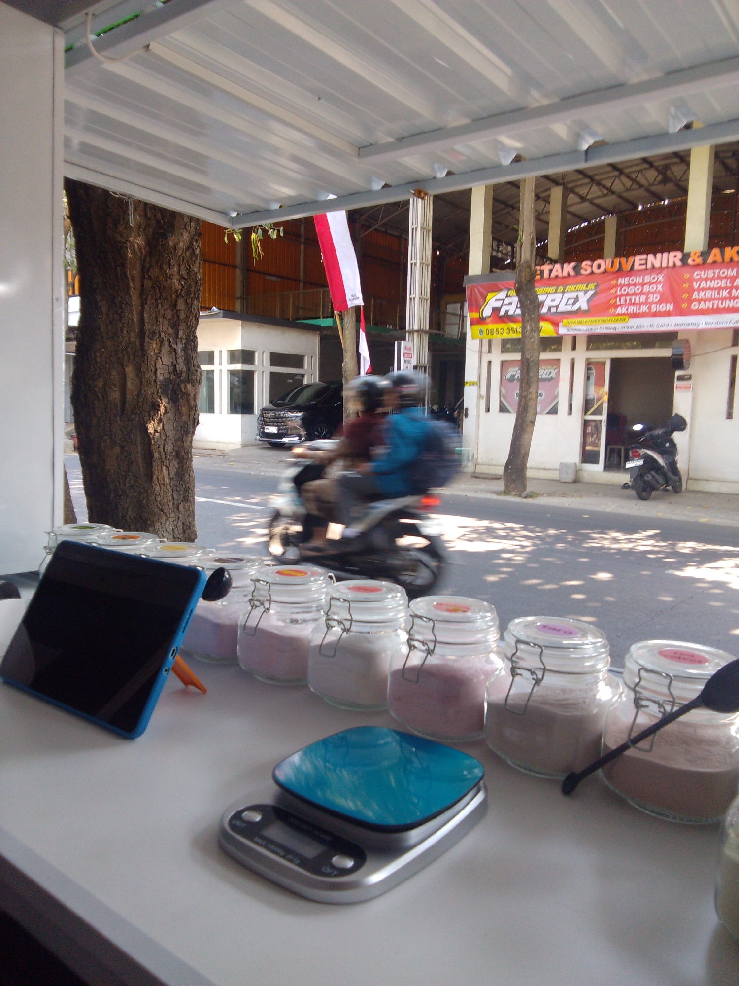 The image depicts a roadside stall in Trenggalek, Indonesia, likely operating under a canopy. In the foreground, on a white surface, are several glass jars containing colored powders, possibly spices or ingredients. A blue tablet computer is propped up on the surface. Next to the jars is a digital scale with a blue tray. In the background, a street scene unfolds with a black car parked on the right and two people on a motorcycle in motion on the left, indicating traffic. A building with signage that appears to be advertisements for printing and souvenir services is visible behind the street. The environment suggests a commercial area with businesses and street activity.