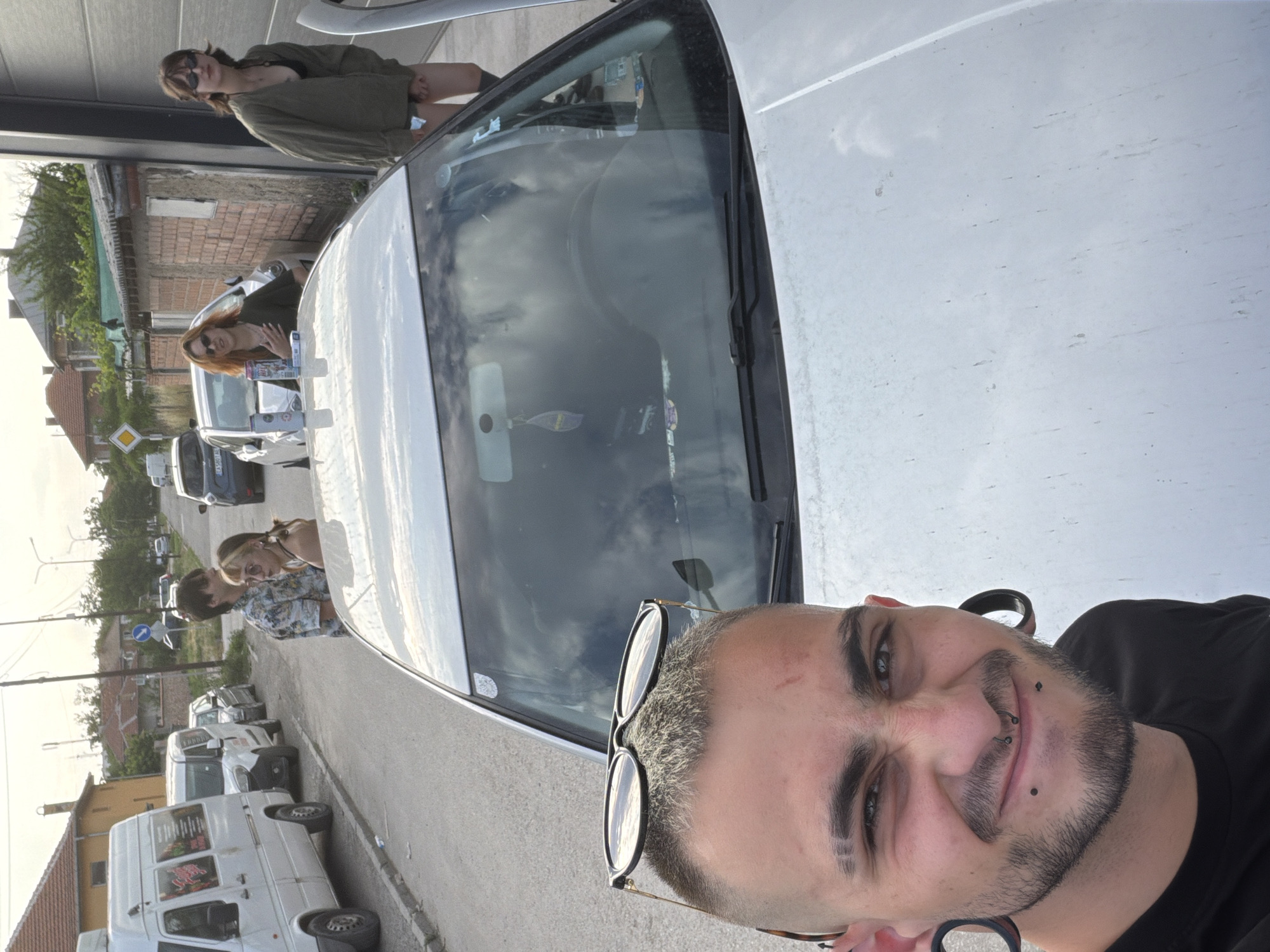 This image captures a street scene in Kalekovets, Bulgaria, at what appears to be late afternoon or early evening under a bright, possibly overcast, sky.
In the immediate foreground, a male with short, light-colored hair and dark shaved eyebrows (one with two shaved lines) is taking a selfie, smiling directly at the camera. He has a short beard, a septum piercing, two lower lip piercings, and large ear gauges. Sunglasses are perched on top of his head, and he wears a black t-shirt. The photo is taken from a low vantage point, looking over the hood of a white hatchback vehicle.
On the white hatchback's hood, near the windshield, a blue, white, and red Pepsi Max can and a small white rectangular box are placed. Inside the car, reflected on the windshield, a purple air freshener hangs from the rearview mirror.
In the mid-ground, a group of four individuals stands around the white hatchback and a nearby brick building. From left to right: a male with dark hair wearing a patterned short-sleeved shirt stands next to a female with light-colored hair, wearing sunglasses and a light top, looking towards the left. Behind the car's hood, a female with reddish-brown hair, wearing dark sunglasses and a dark green long-sleeved top, leans against the vehicle, her mouth open. To her right, a fourth female with dark, shoulder-length hair, wearing dark sunglasses, a dark green jacket over a dark top, dark shorts, and high socks, stands beside a light-colored building with a large dark opening, potentially resting an arm on an open car door.
Further down the street, a white Mercedes-Benz Sprinter-style van with colorful decals is parked on the left side, followed by a smaller white van. Several other vehicles, including a dark blue/grey hatchback, are visible parked or driving in the background.
The street itself is paved with asphalt, featuring curbs and visible debris along the roadside. Residential buildings with terracotta-tiled roofs are visible in the background, along with a prominent brick wall on the right. Utility poles and overhead lines are present, and a yellow diamond "priority road" sign is visible in the mid-background.