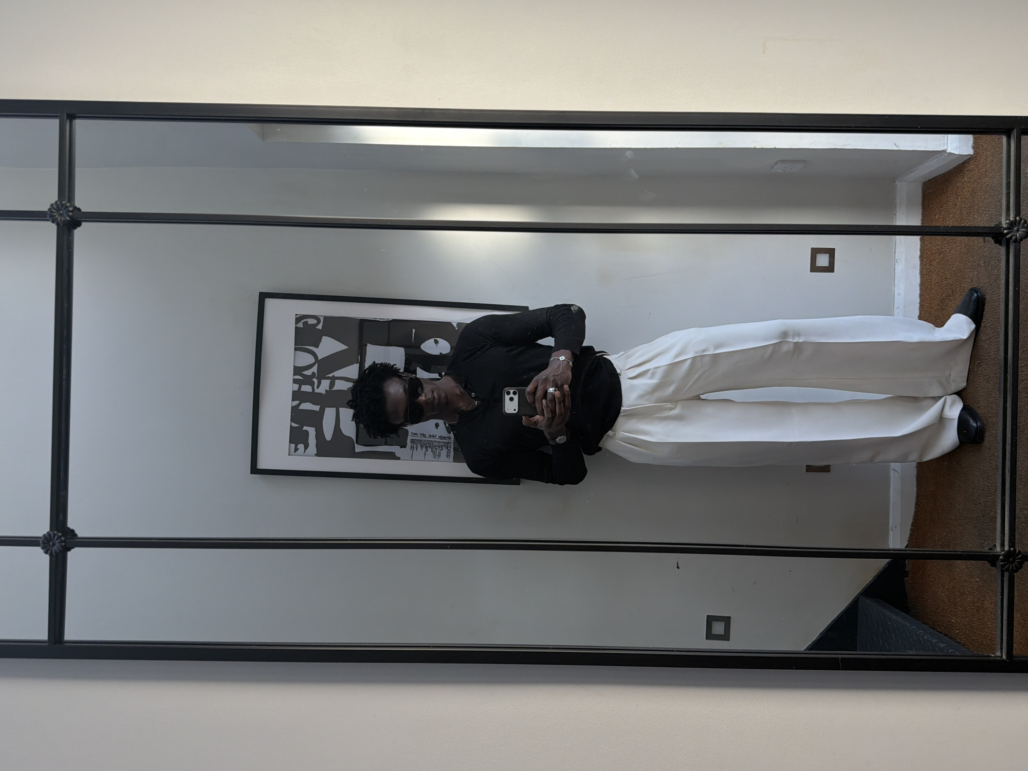 A mirror selfie captures a person with dark hair and sunglasses, wearing a black top and white trousers, holding a mobile phone. Reflected in the mirror is a black-and-white framed poster on the wall. The room appears to be indoors and partially shows an electrical outlet and the edge of a brown carpet. The mirror has a decorative black metal frame. There is no evidence to confirm that the location is London, United Kingdom.