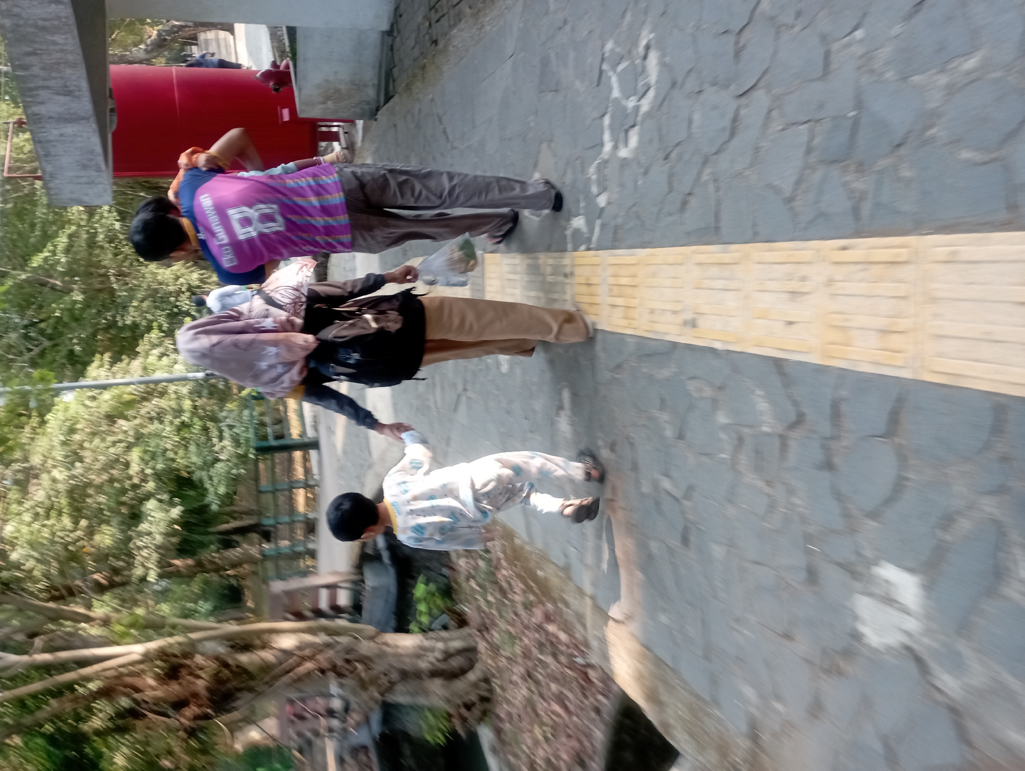 In Depok, Indonesia, a family walks along a textured pathway. A man carries a young child on his shoulders, while a woman walks beside them. Another young child, dressed in pajamas, walks ahead, holding the hand of the woman. The path is bordered by trees and a red structure.