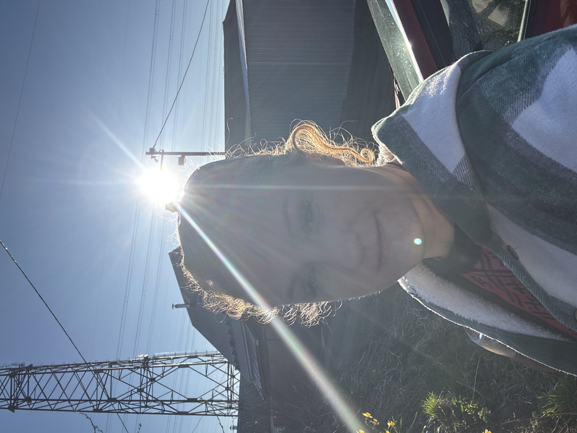 A bright, sunlit outdoor scene is captured in a sideways orientation. The primary subject is a person, likely female, whose upper body and face are partially obscured by a strong sun flare emanating from the top left of the frame. She has curly, light-colored hair, with individual strands illuminated by the backlighting. Her visible eye shows a slight smile. She is wearing a green and white plaid jacket, possibly lined with a white sherpa-like material, and a patterned red garment is visible underneath.
The background features a clear blue sky, traversed by numerous power lines. Large, dark grey industrial structures, potentially part of a power plant or industrial complex, are visible behind the person, extending towards the bottom of the frame. A prominent metal electricity pylon or tower structure is visible in the lower left, characterized by its crisscrossed supports. Part of a red vehicle, likely a car door or frame, is visible on the far right, suggesting the person is in or leaning out of a vehicle. In the bottom right corner, sparse green vegetation and small yellow flowers are visible on the ground.
The strong sun indicates a daytime setting, possibly morning or late afternoon given its low angle. The presence of extensive power lines, pylons, and industrial buildings suggests an infrastructure-heavy area, consistent with the provided location context of Chaikovsky, Russia. No text is visible in the image.